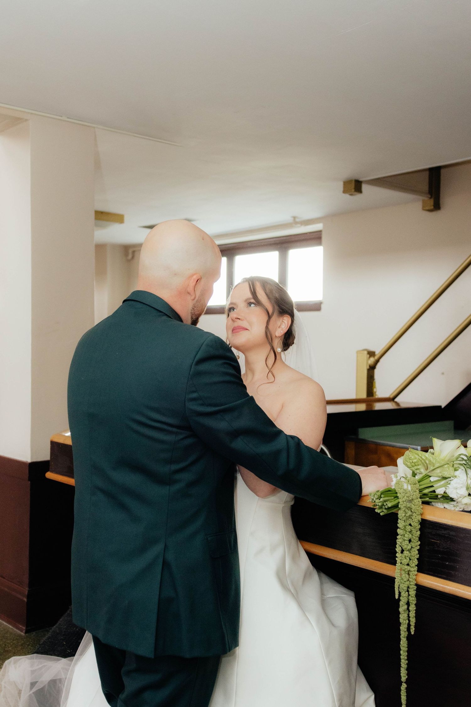 Groom in dark suit holds bride in white strapless gown near staircase.
