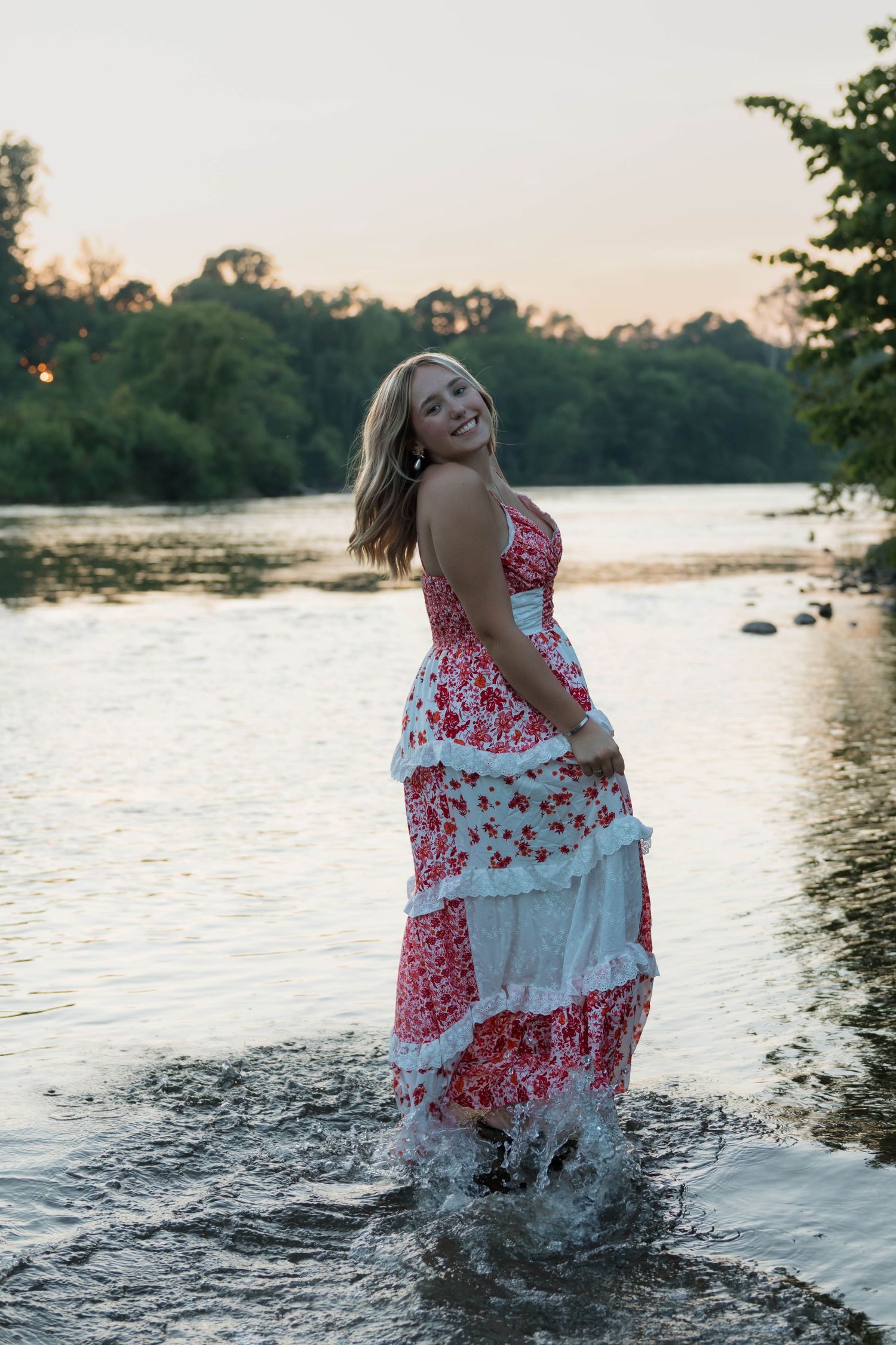Joyful blonde woman in red floral dress splashing in river at sunset during senior photo session.