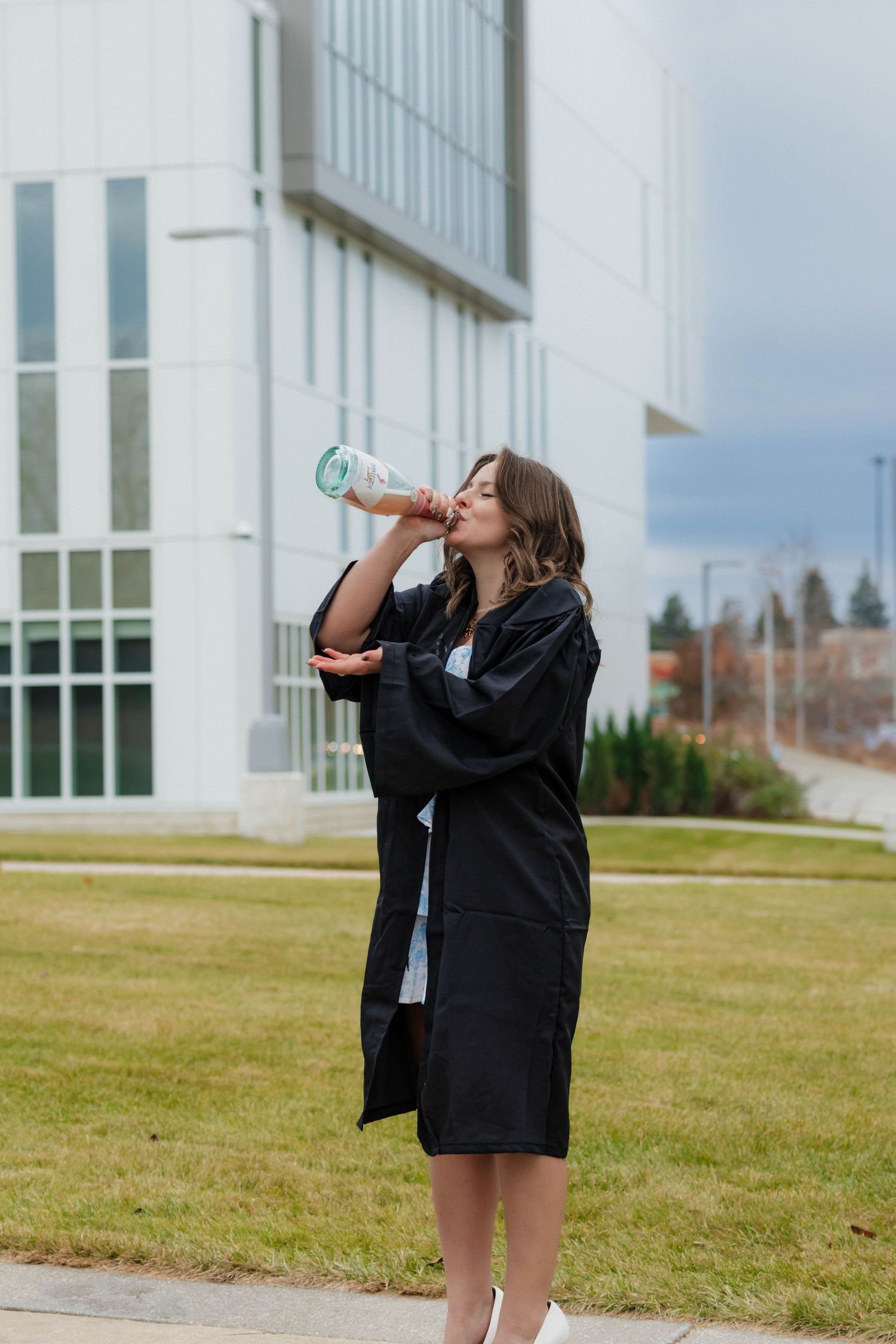 Young woman in graduation gown drinking from a water bottle while standing on a green campus lawn.