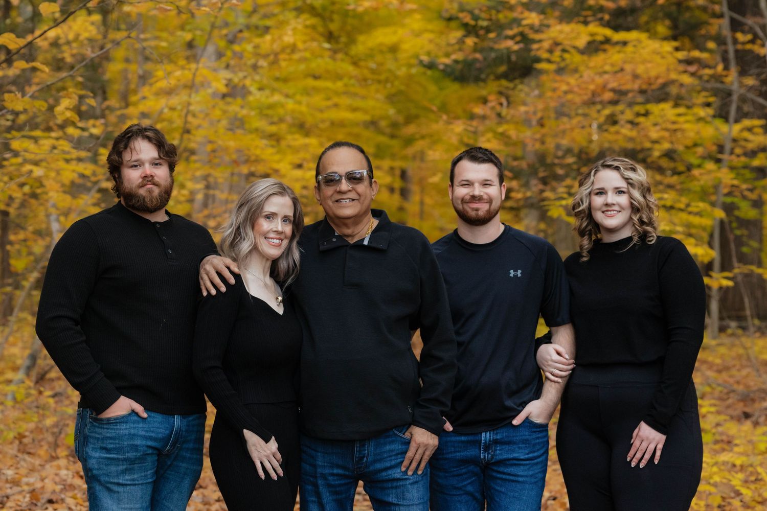 Family of five posing in matching black outfits among vibrant autumn foliage in a forest setting.