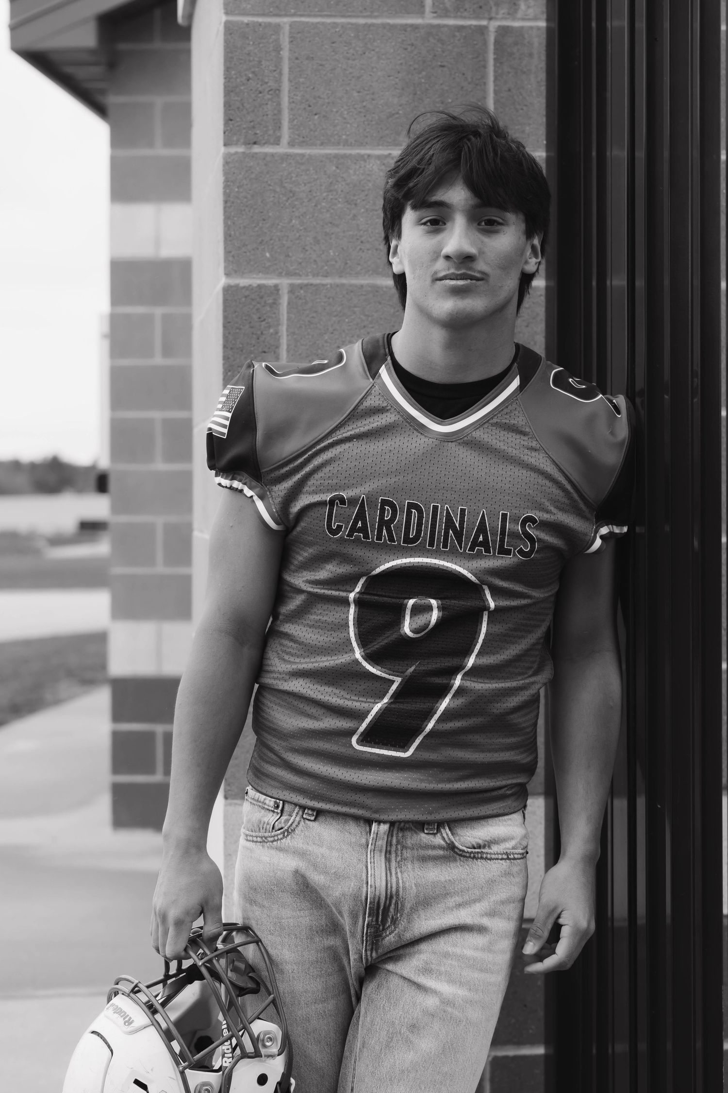Black and white portrait of teen in Cardinals #9 football jersey holding helmet outdoors.