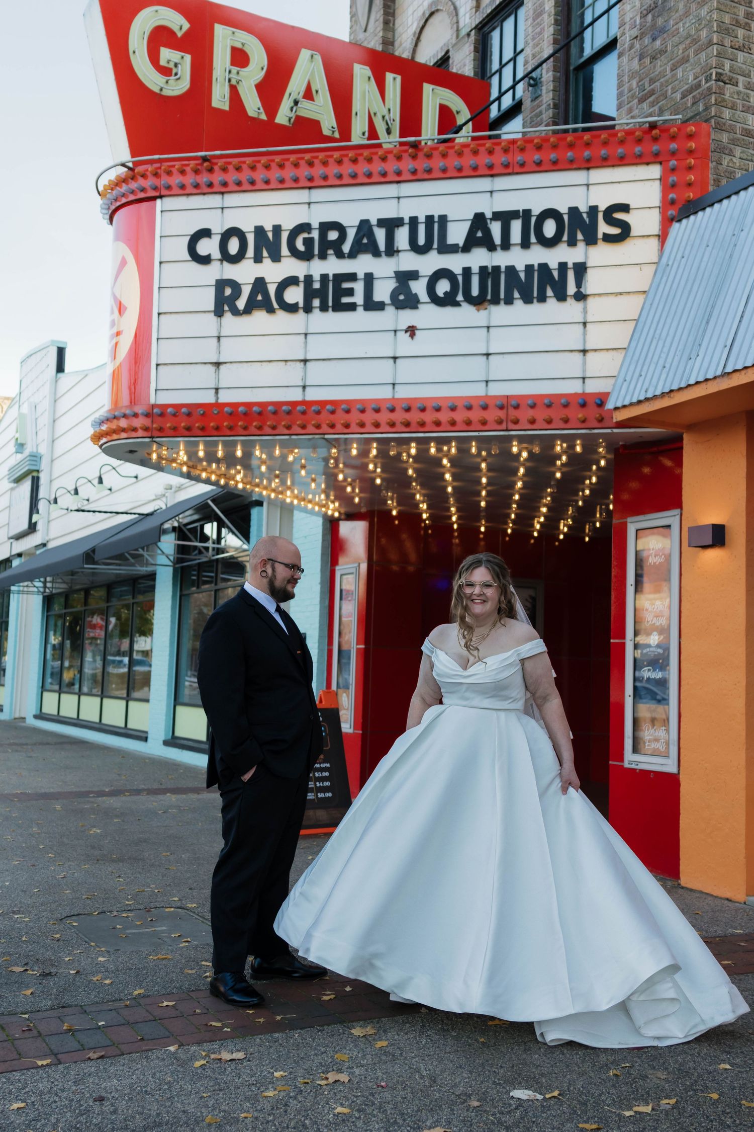 Bride twirls white ballgown while groom watches under lit Grand Theatre wedding congratulations marquee sign.