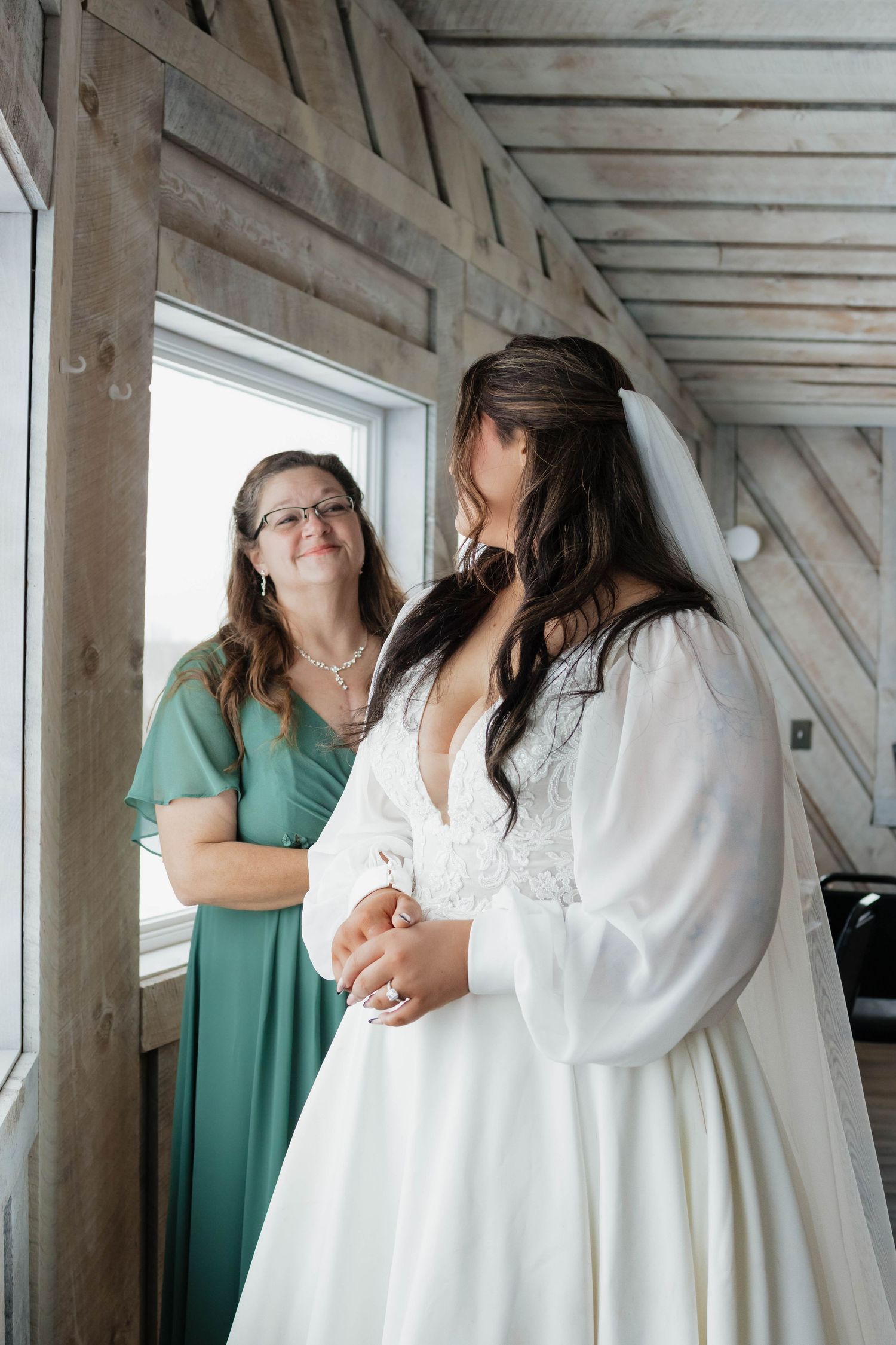 Bridesmaid in green dress helping bride in white lace gown prepare near rustic wooden window.