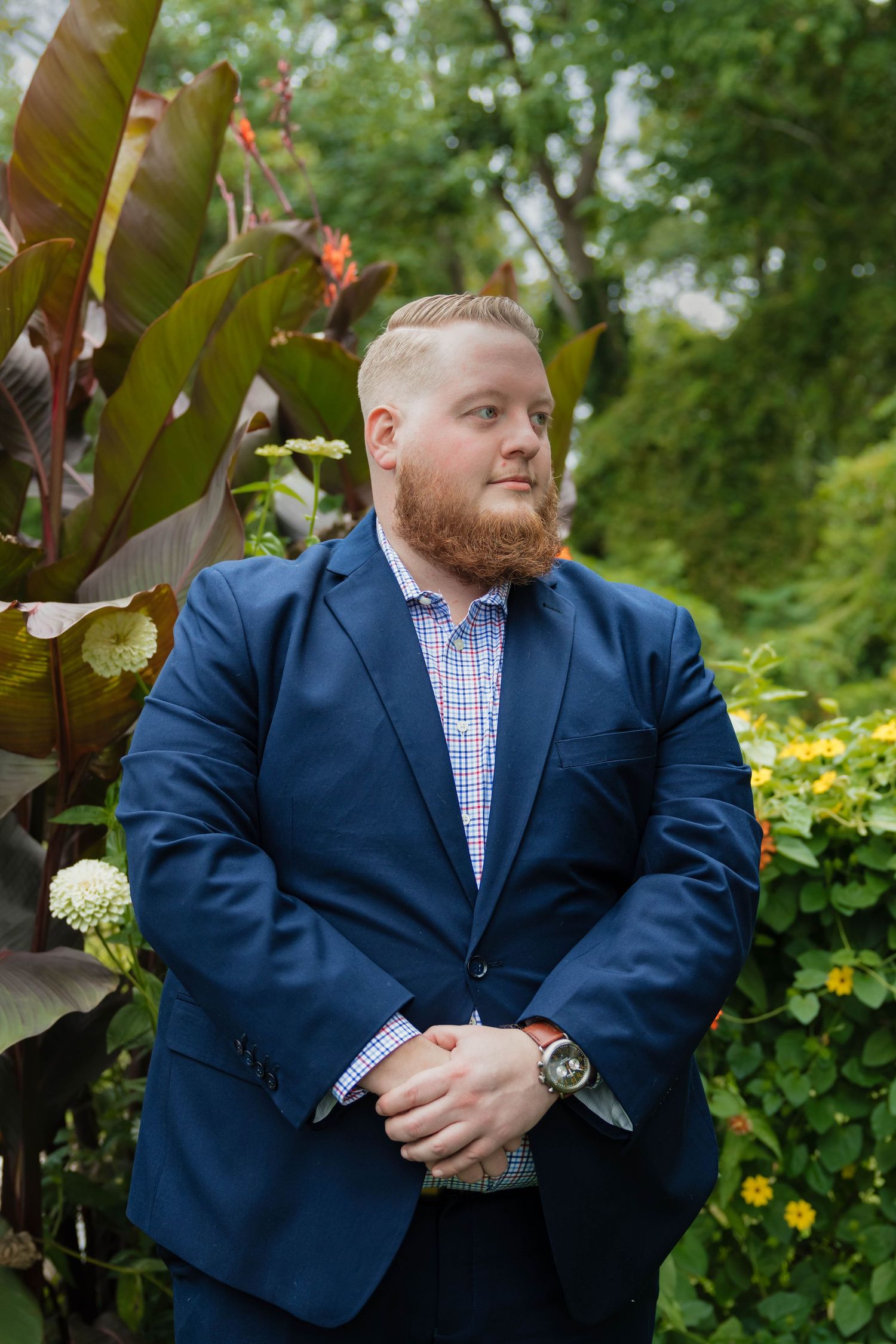 Bearded redhead man in navy blazer and plaid shirt poses confidently in lush garden setting.