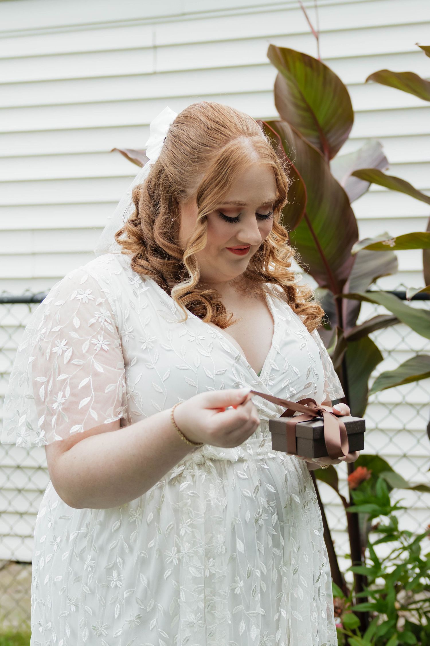 Bride in white lace gown opens a small gift box outdoors near tropical plants.