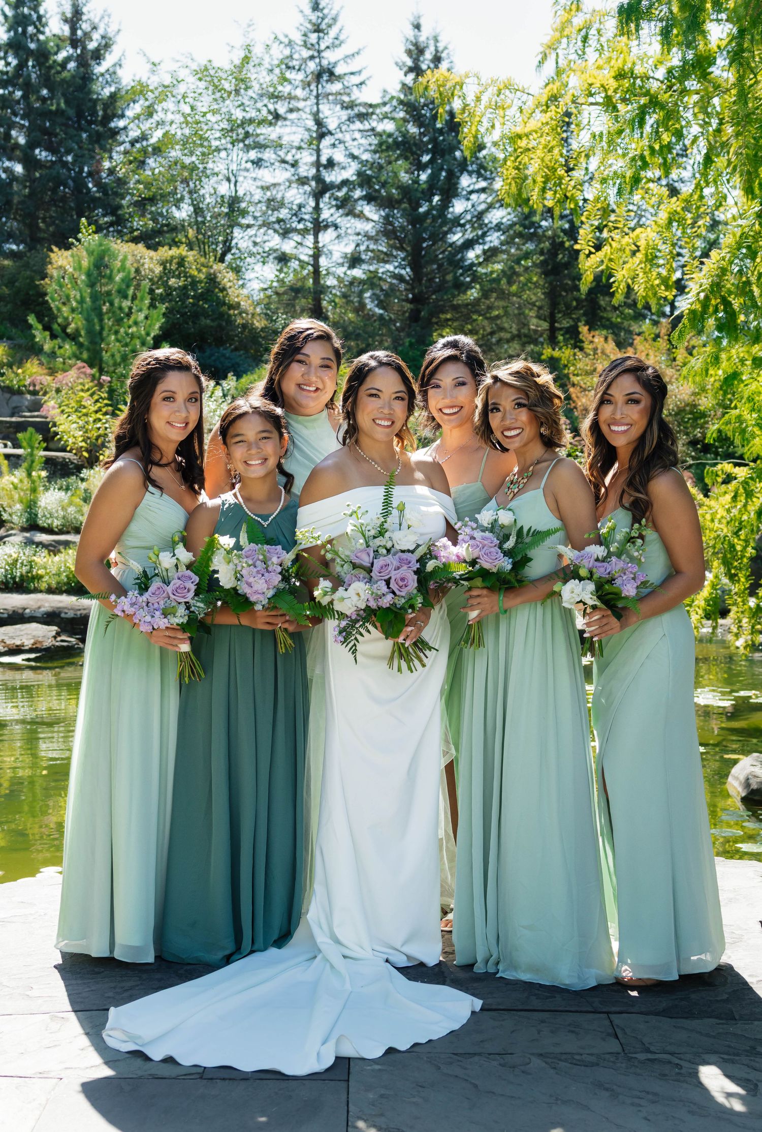 Bride and five bridesmaids in sage green gowns smile holding lavender bouquets beside a sunlit garden pond.