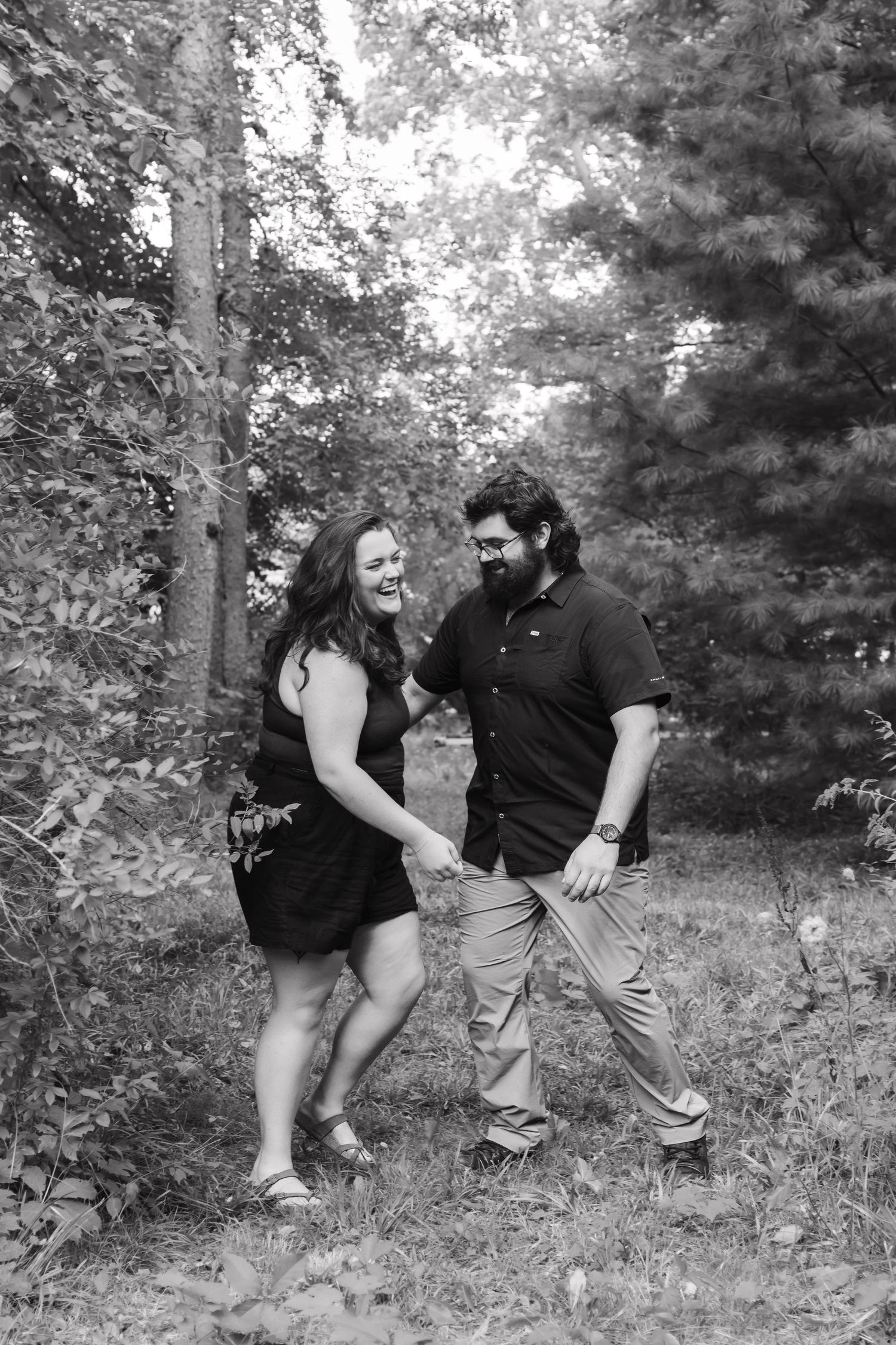 Black and white photo of a happy couple holding hands and laughing while walking through a lush forest.