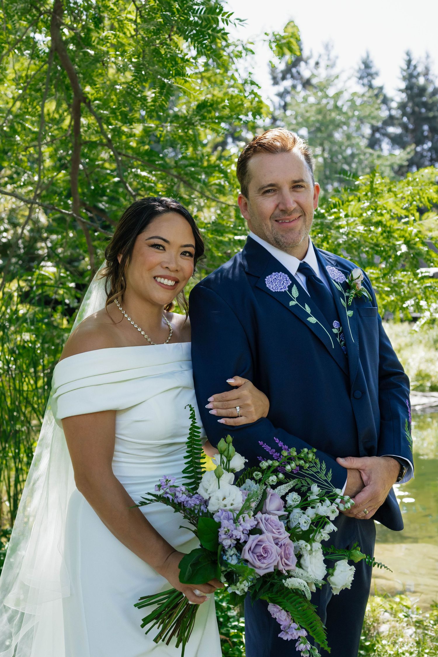 Bride in off-shoulder white gown and groom in navy suit posing with purple floral bouquet.