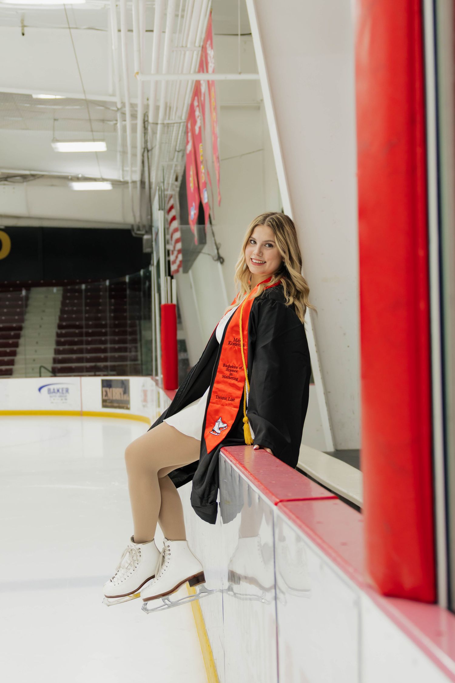 Graduate in black gown and orange stole sits on hockey rink boards wearing white ice skates.