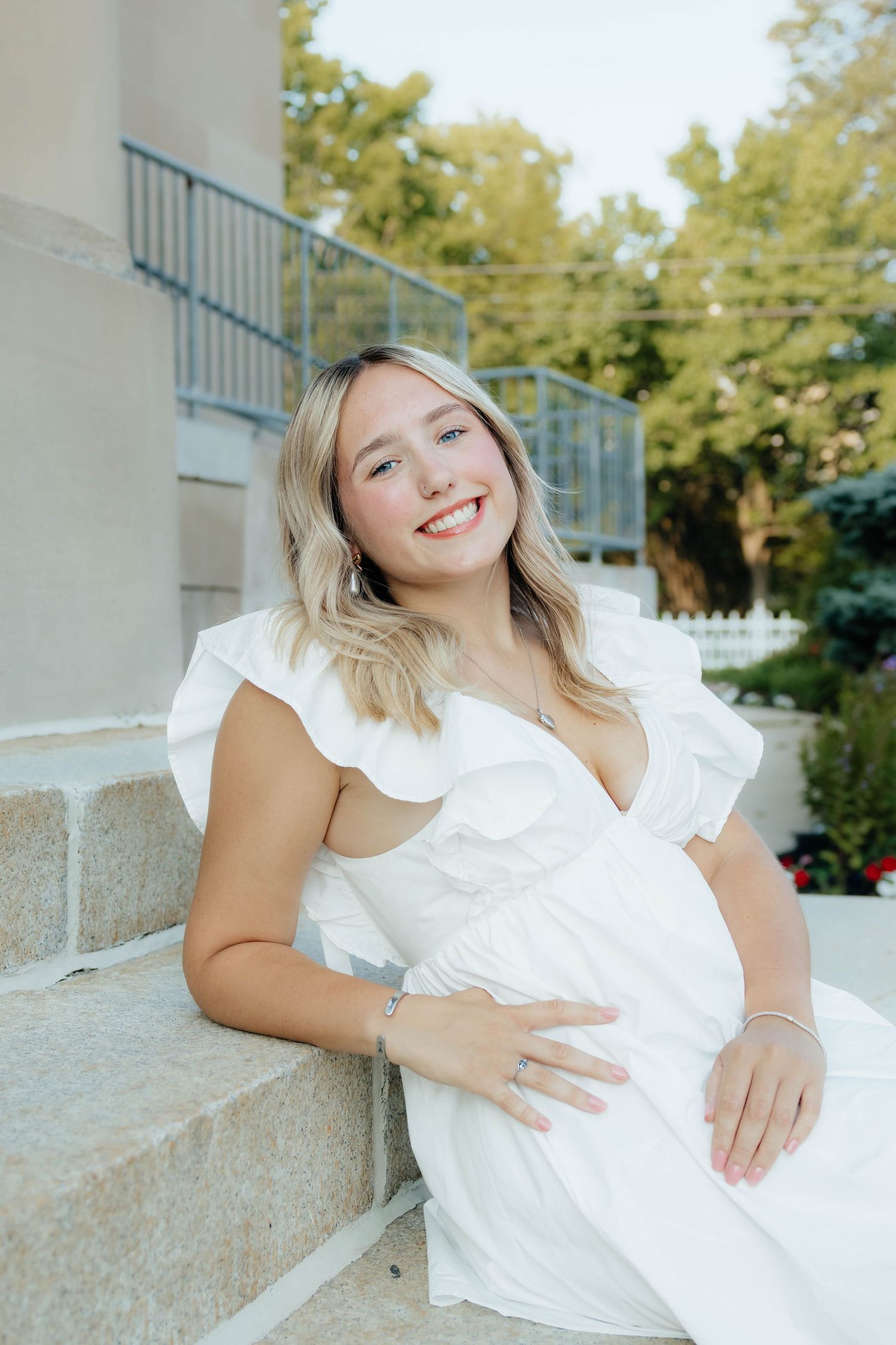 Smiling pregnant woman in white ruffled dress sitting on stone steps outdoors.