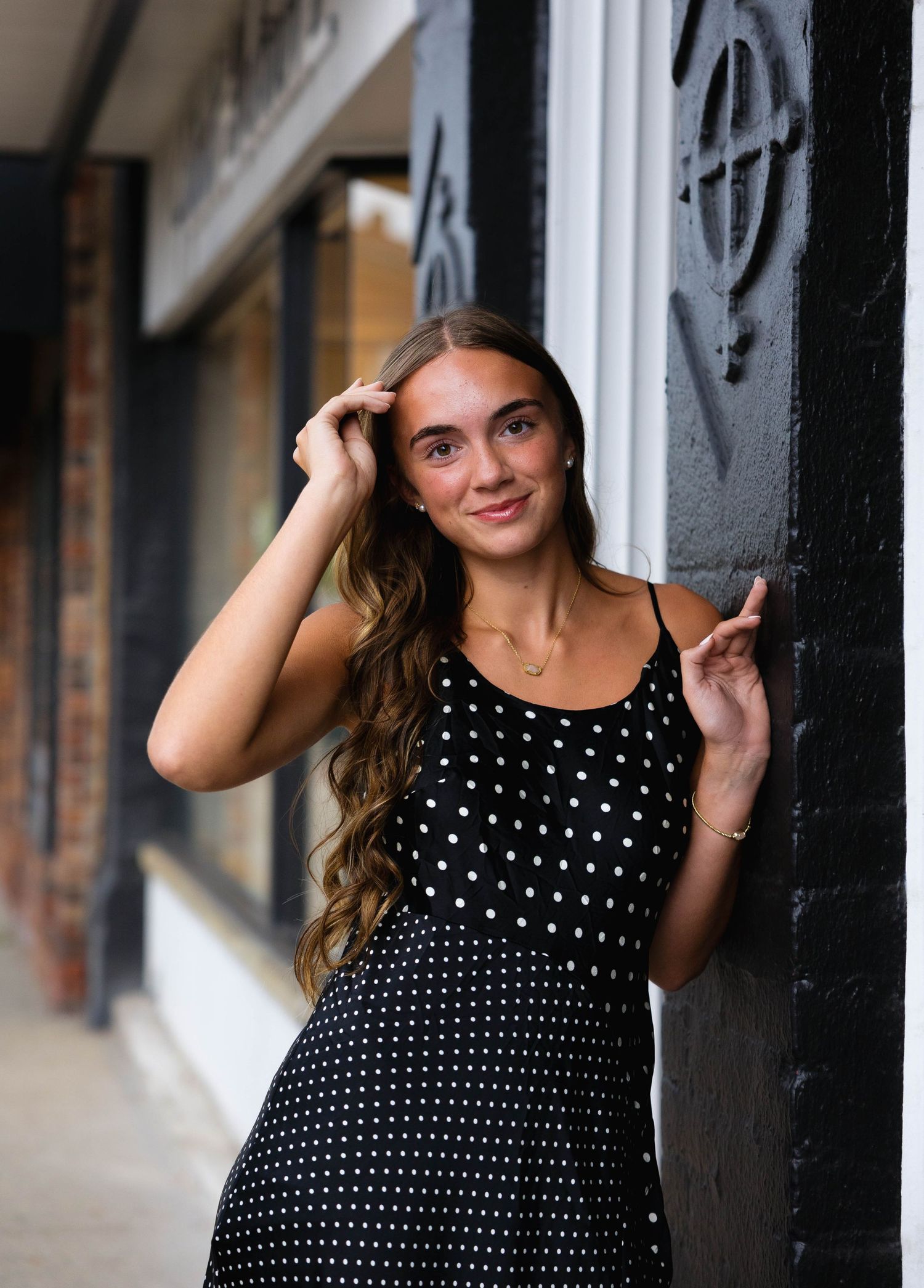 Young woman in black polka dot dress posing against decorative pillar on urban street.