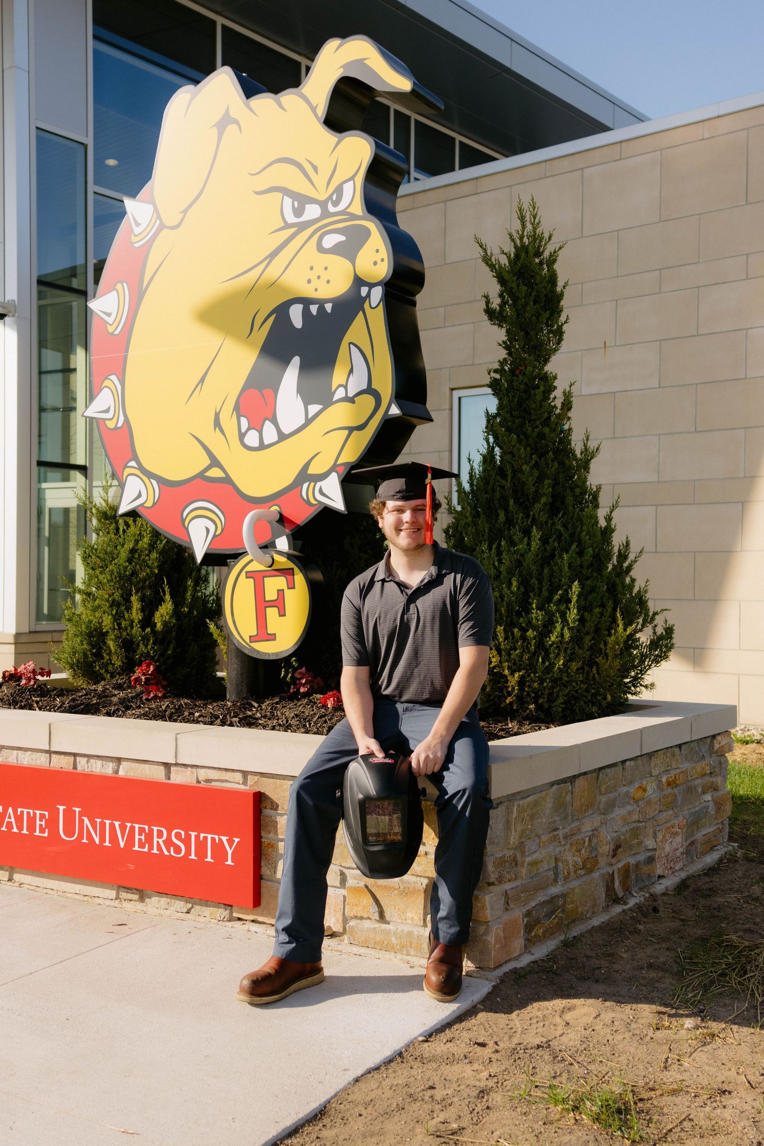Young male graduate sits in front of Ferris State University Bulldog mascot sign wearing graduation cap outdoors.
