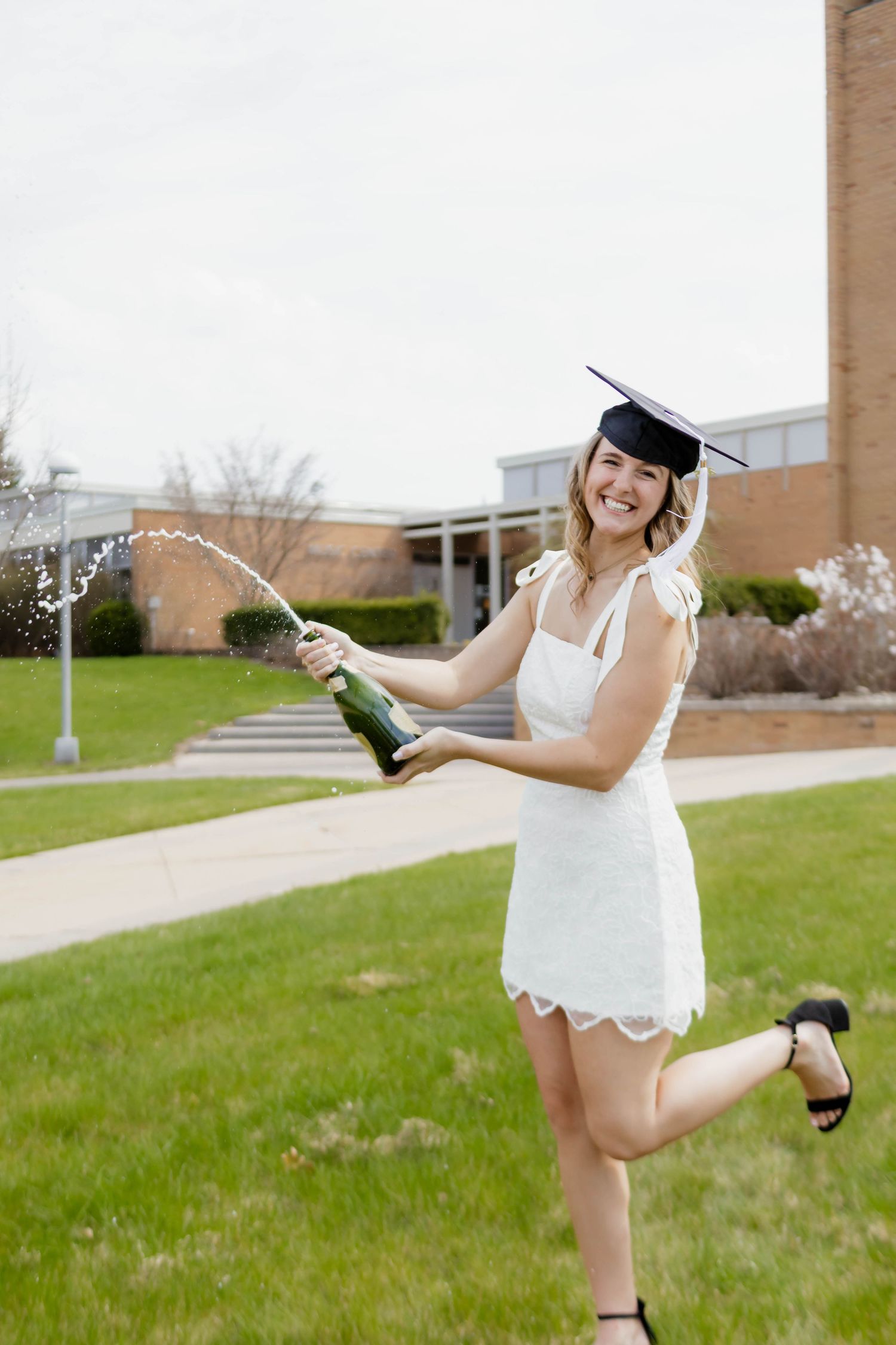 Smiling female graduate in white dress and cap pops champagne bottle on college campus lawn.