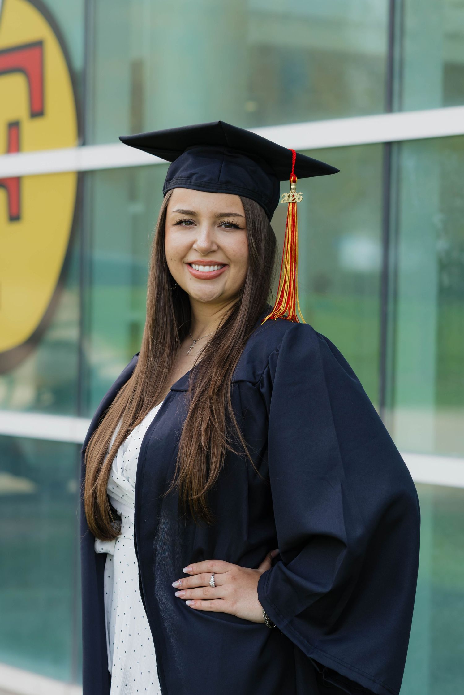 Smiling graduate in navy cap and gown with red and gold tassel poses before glass building.
