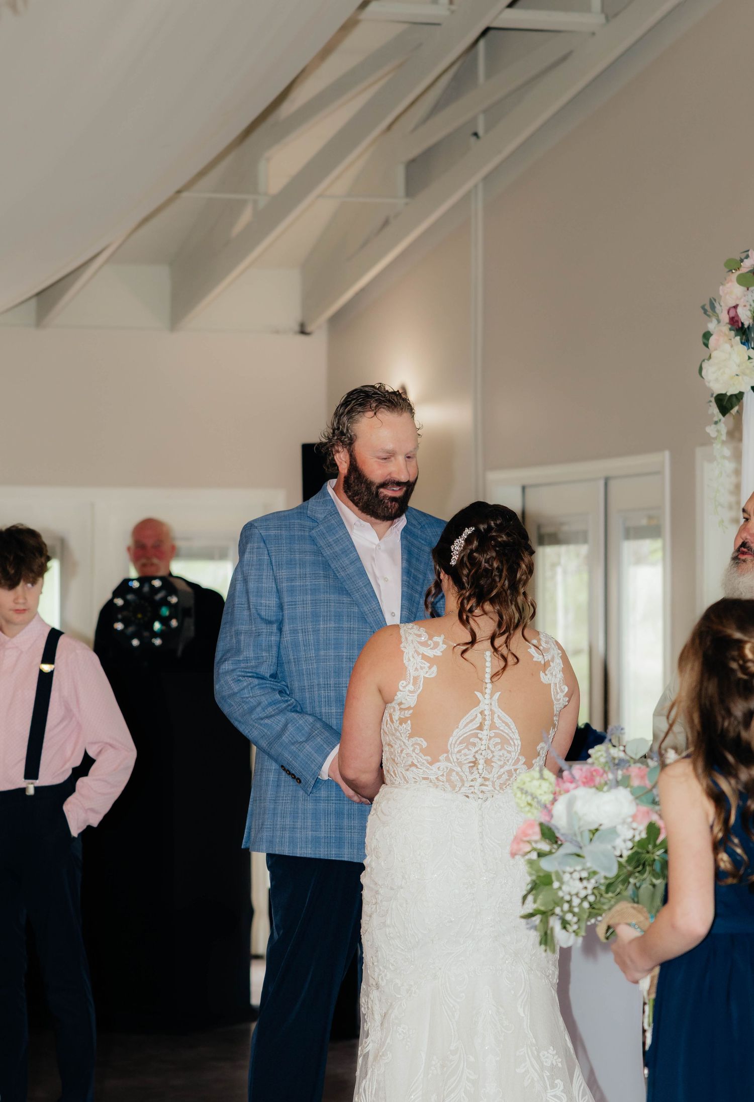 Bride in lace gown and groom in blue plaid suit share a tender moment during indoor wedding ceremony.