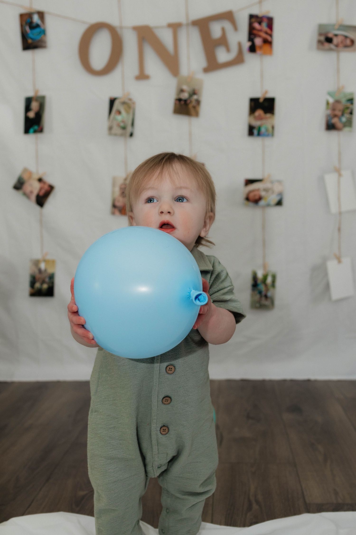 One-year-old boy holds blue balloon standing before photo display wall with ONE banner.