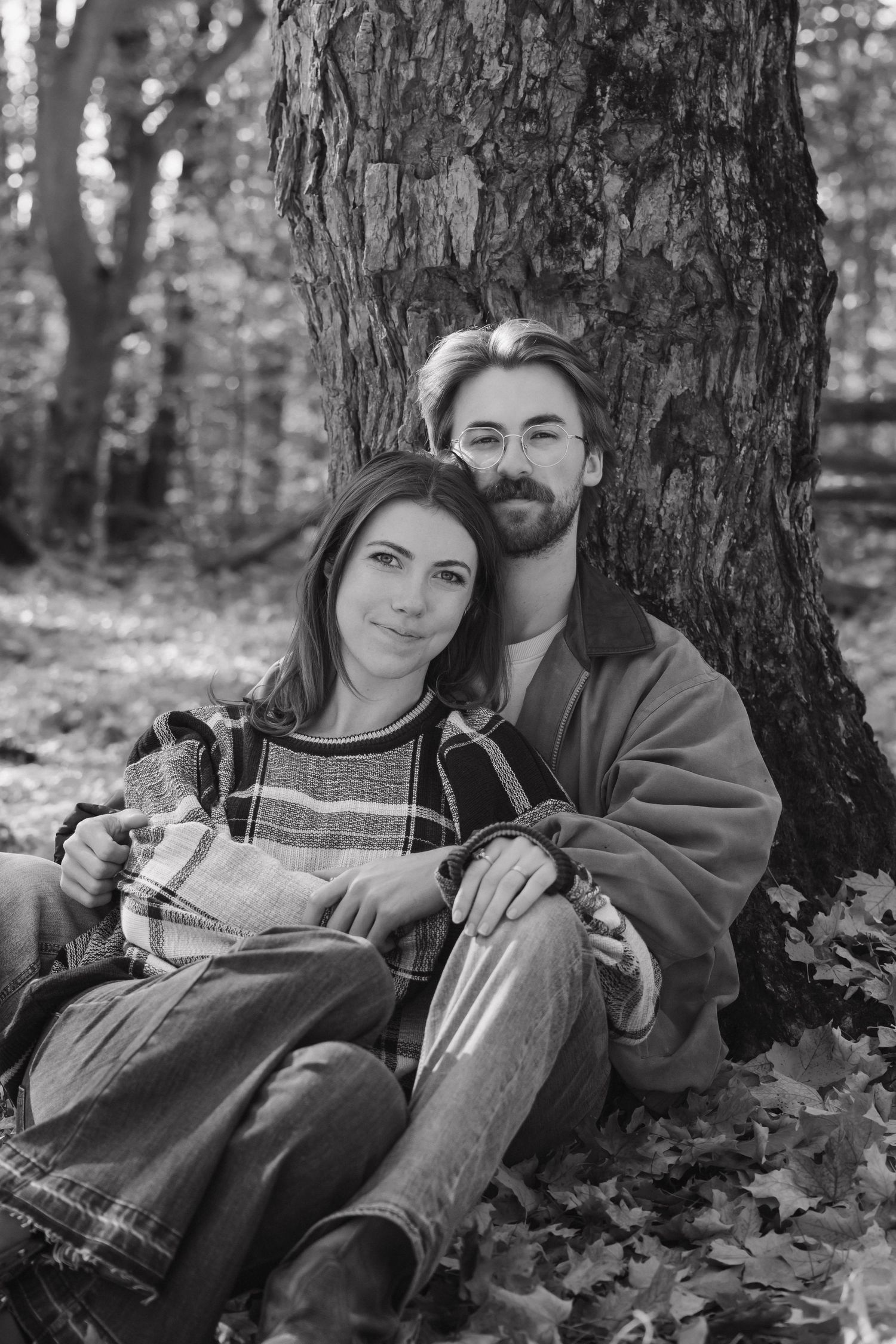 Black and white photo of a couple sitting together leaning against a tree in a forest.