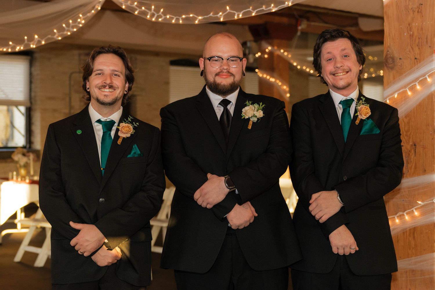 Three groomsmen in black suits with teal boutonnieres smiling at a rustic wedding venue with string lights.