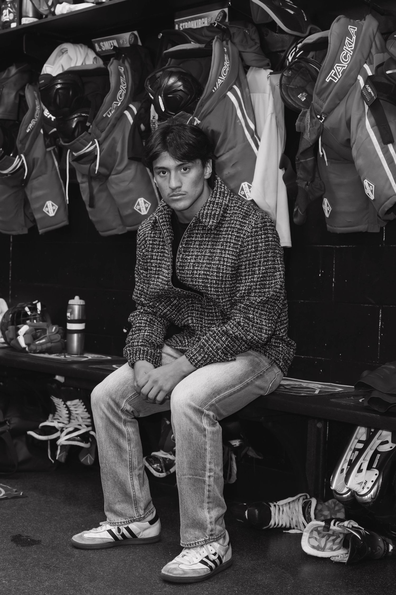 Young man in tweed jacket sits in hockey locker room surrounded by gear, black and white.