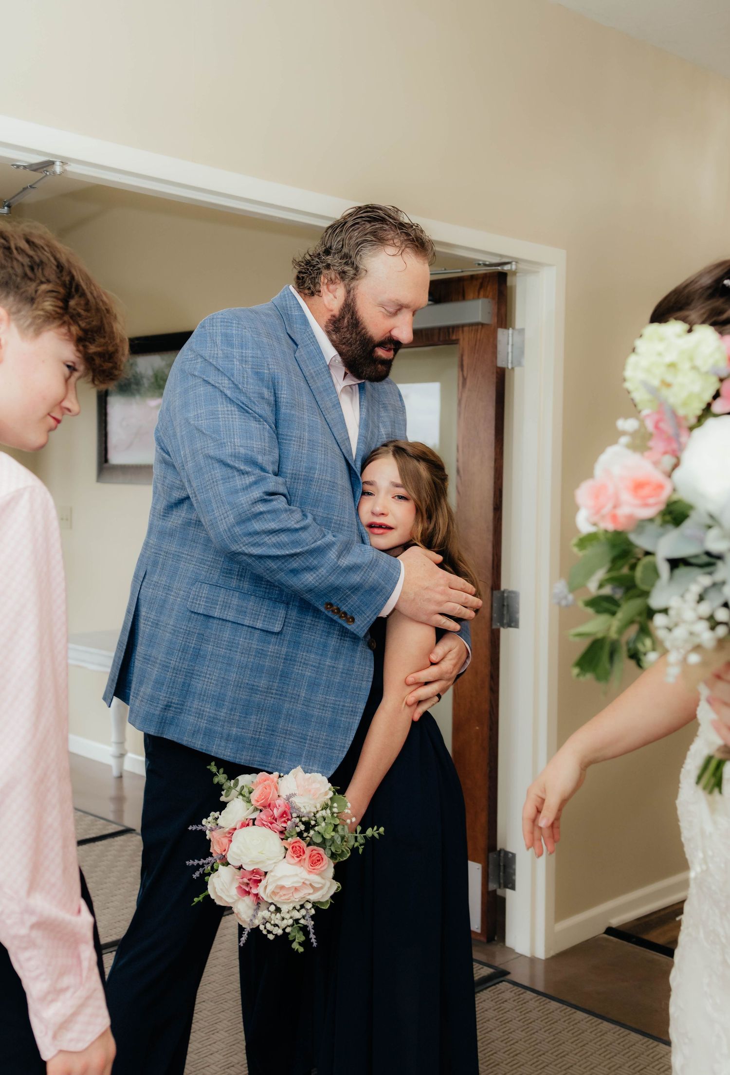 Groom in blue plaid suit embraces emotional flower girl in navy dress during wedding day first look.