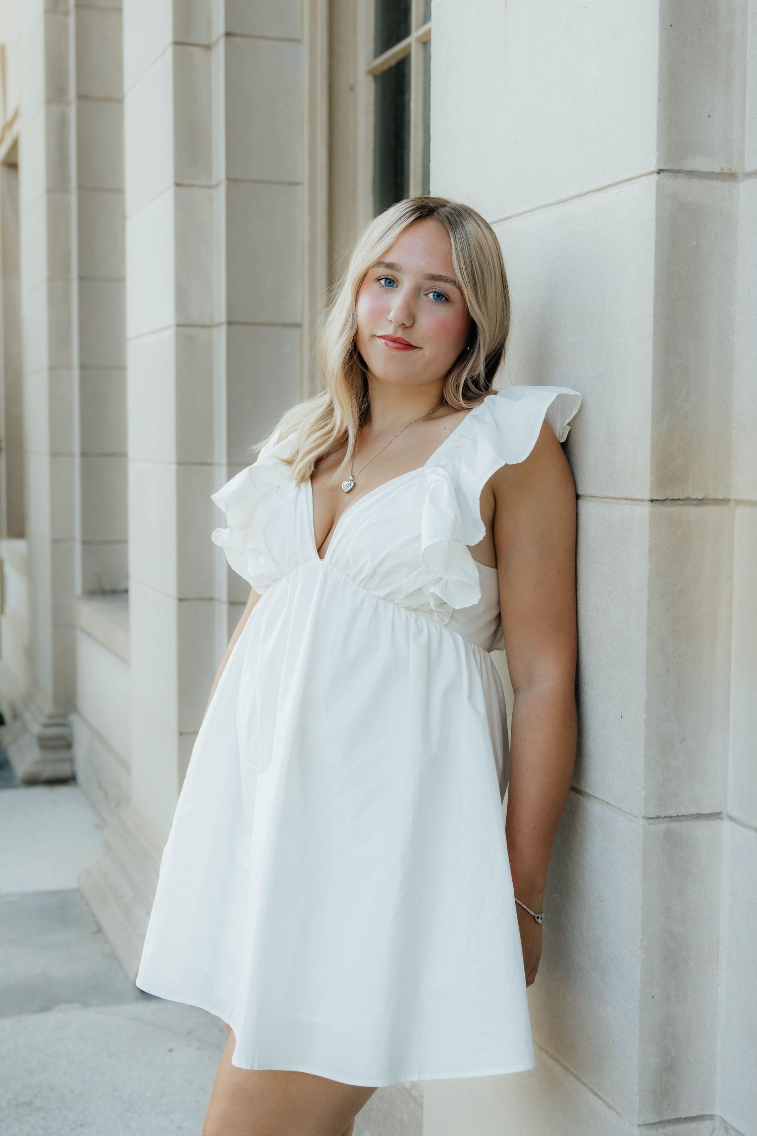 Elegant blonde woman in white ruffled mini dress leaning against a stone column outdoors.