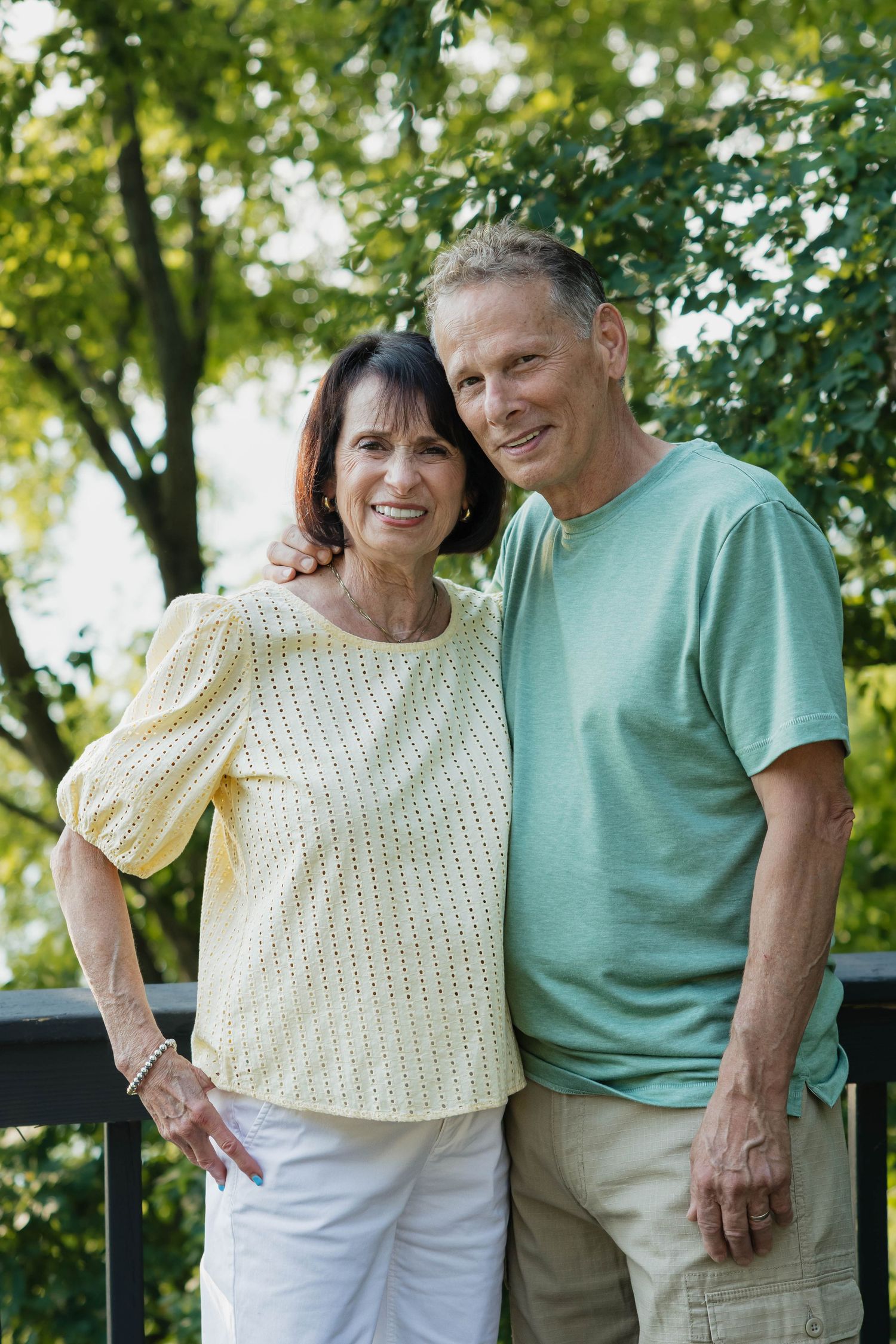 Smiling middle-aged couple posing outdoors on a deck surrounded by lush green trees.