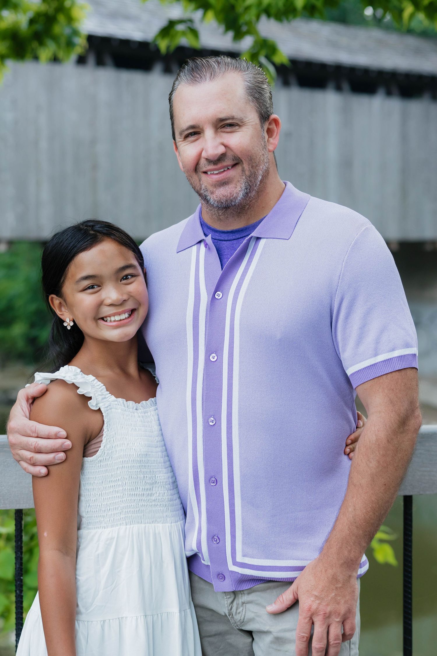 Happy young girl and middle-aged man smile together outdoors near a covered bridge.