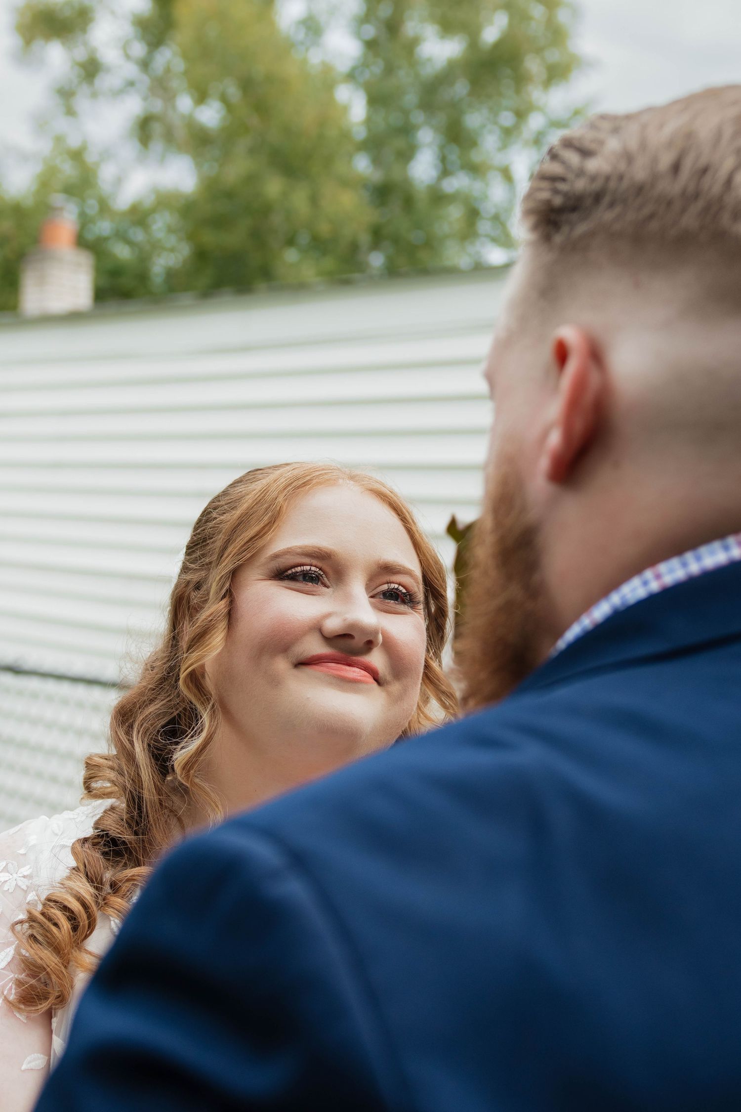 Bride with curly red hair smiling up at bearded groom in navy suit outdoors.