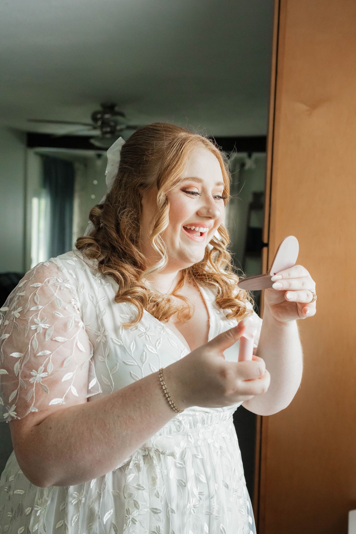 Radiant bride in white lace dress checks her makeup in a small handheld mirror.