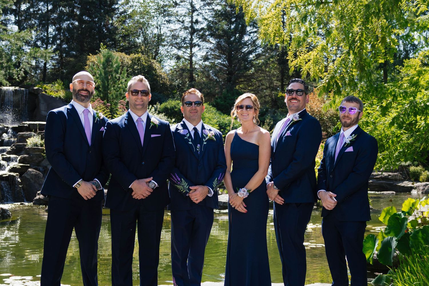 Wedding party in navy suits and blue gown posing outdoors near a pond in a lush garden setting.