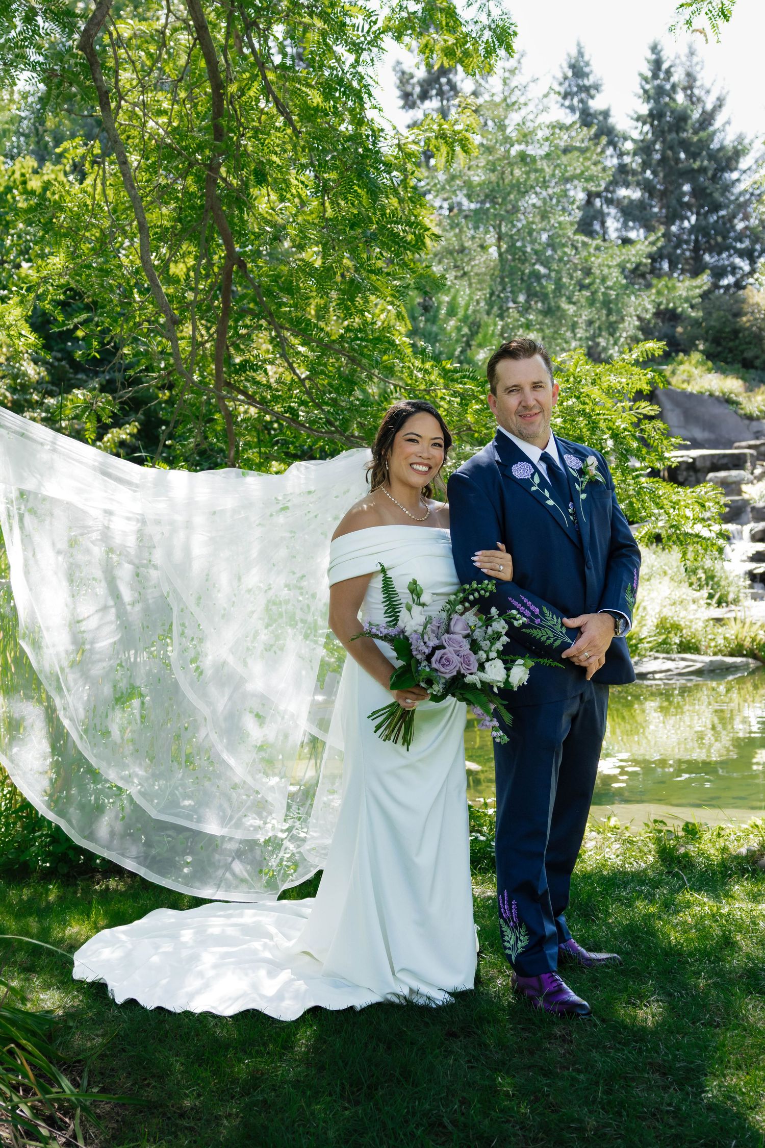 Bride's veil flowing in the wind as couple poses by a garden pond in sunlight.
