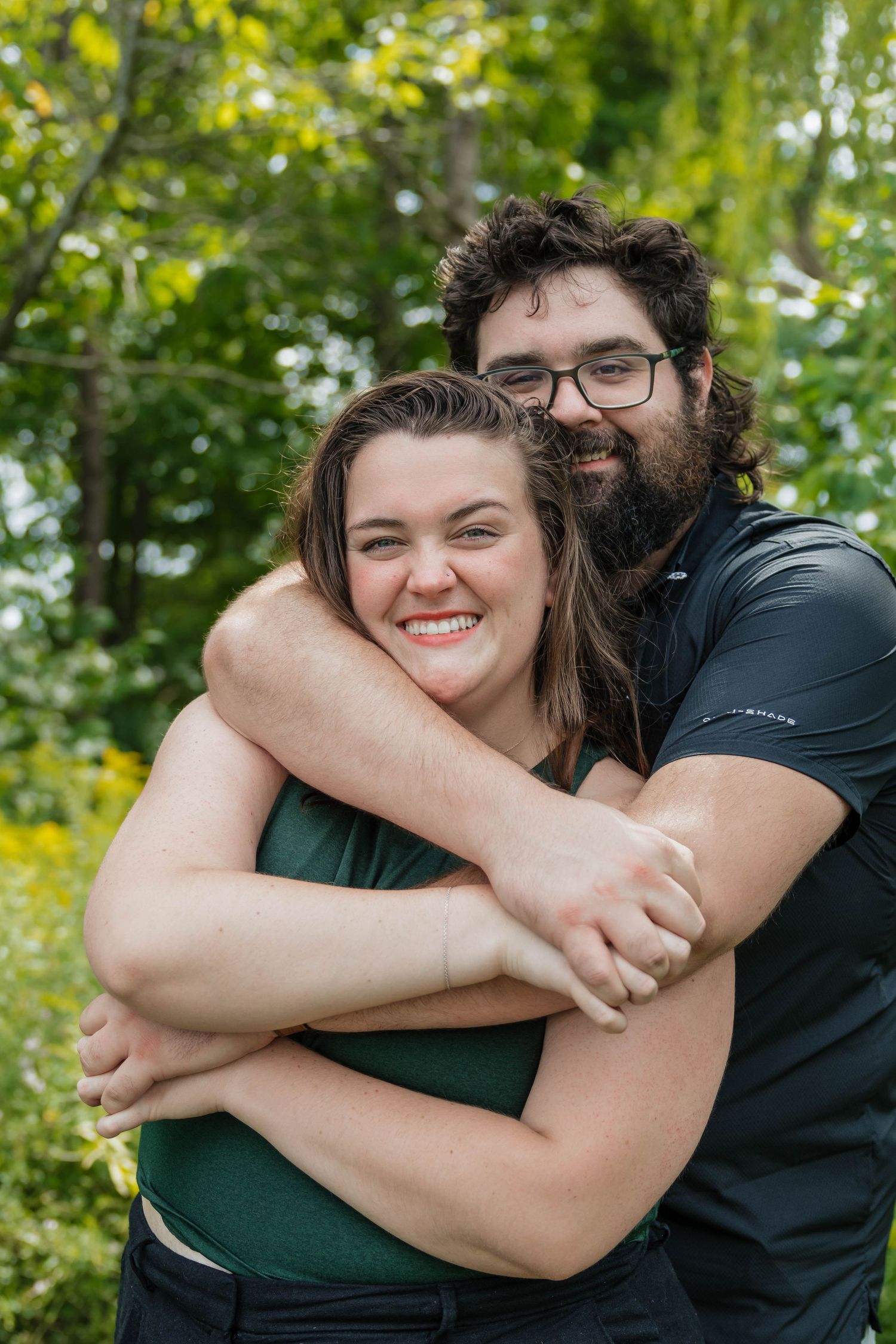 Bearded man in black shirt hugging smiling woman in green top during outdoor engagement photo session.
