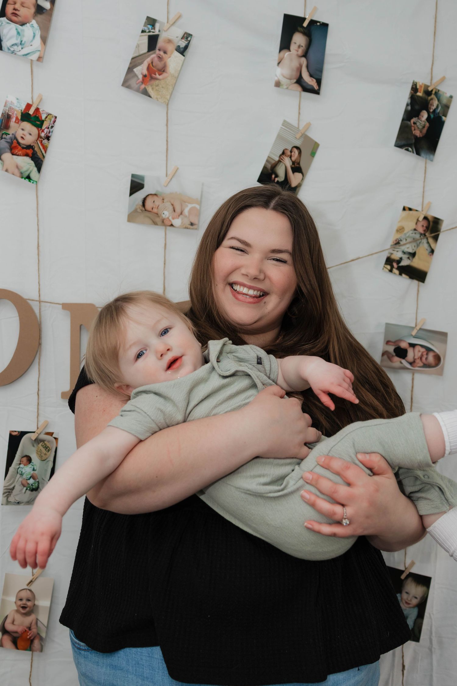 Smiling mother holds laughing one-year-old baby boy in front of birthday photo display wall.