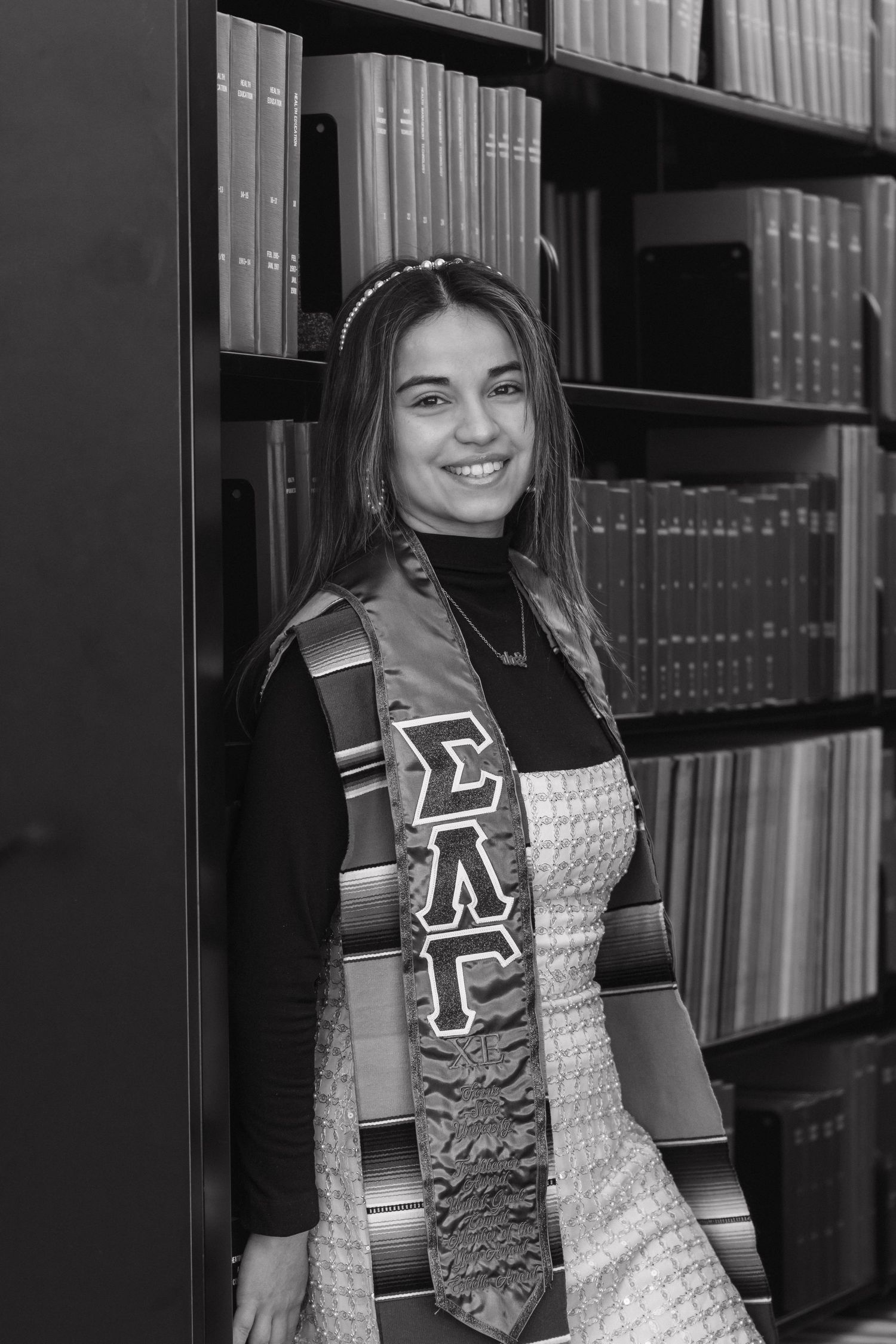 Black and white photo of graduate wearing Sigma Lambda Gamma stole standing before library bookshelves.