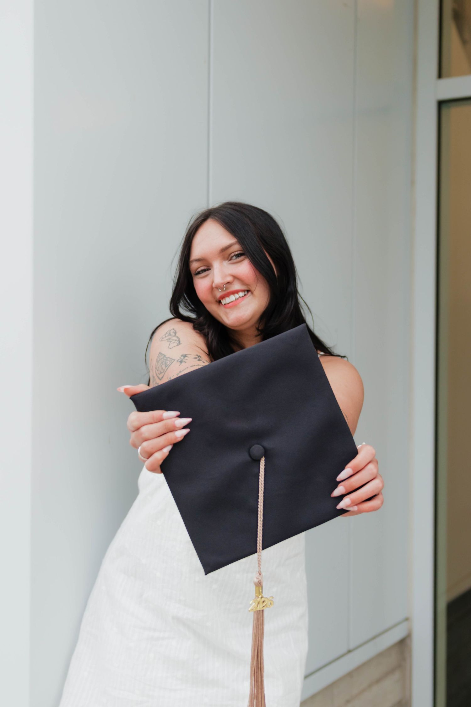 Smiling brunette graduate in white dress hugging black mortarboard cap with gold tassel close to her face.
