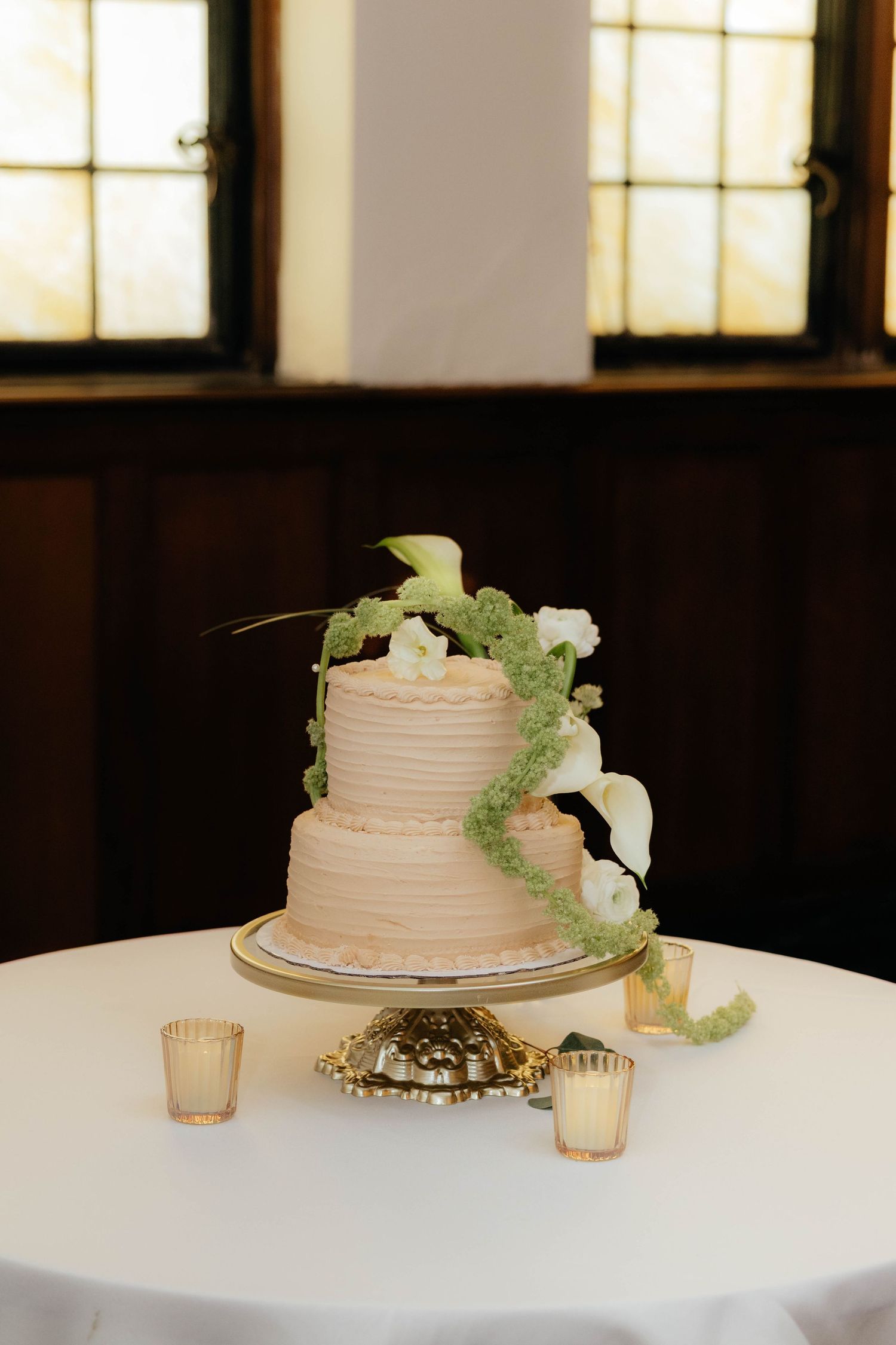 Two-tier blush wedding cake with white calla lilies and green floral accents on gold cake stand.