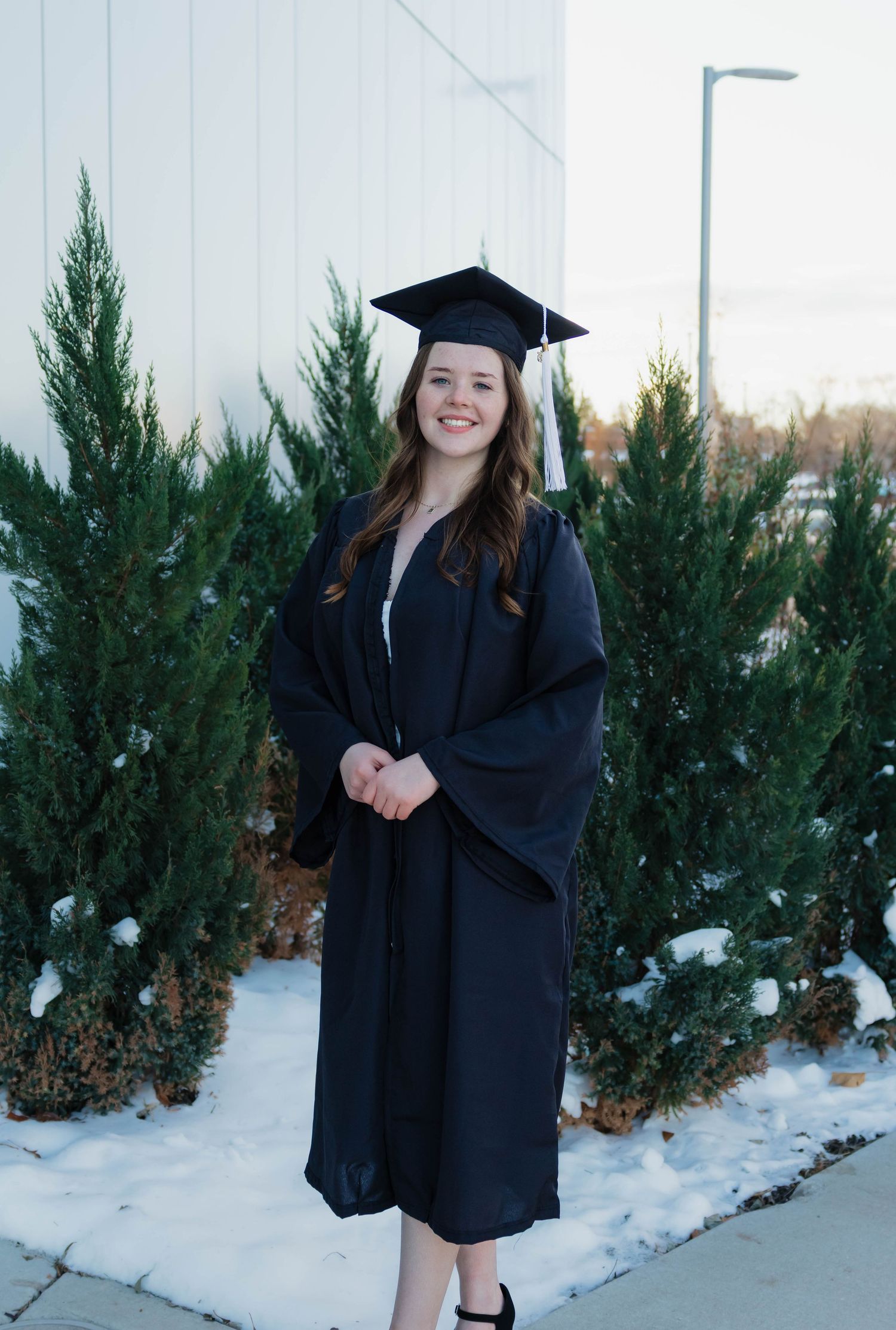 Young woman smiling in black graduation cap and gown standing outdoors in winter with snow on ground.