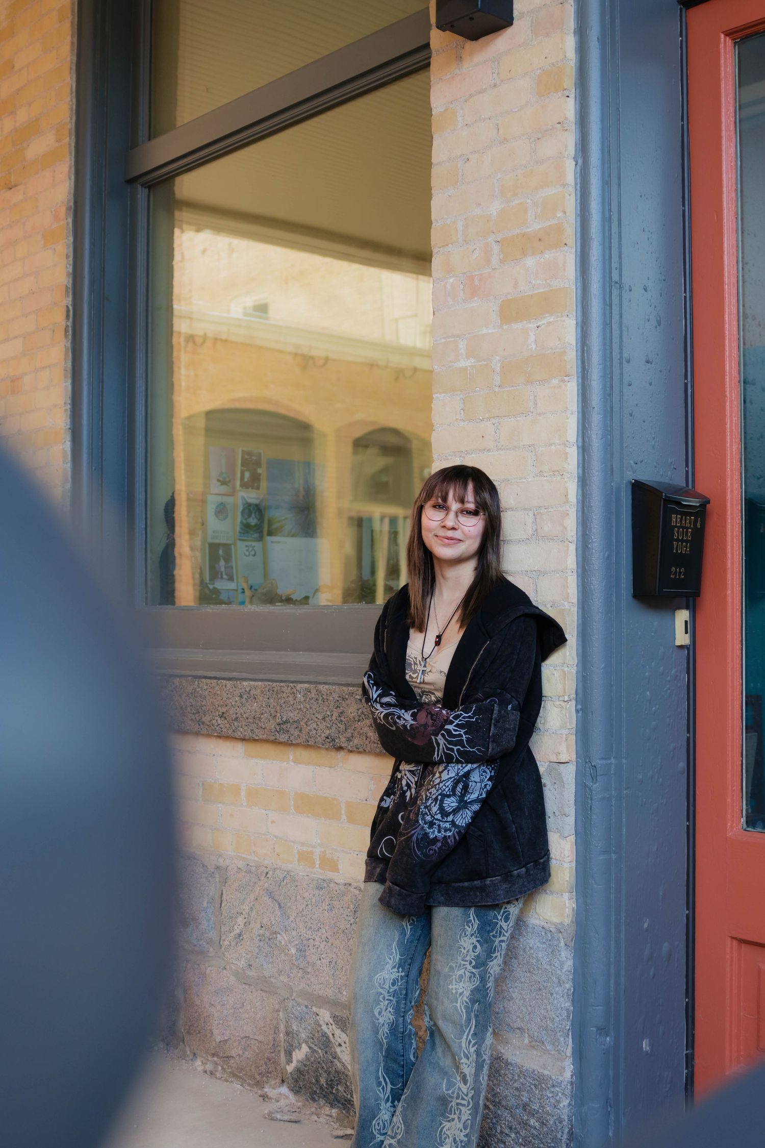 Young woman with bangs and glasses leaning against a brick building doorway in casual street style outfit.