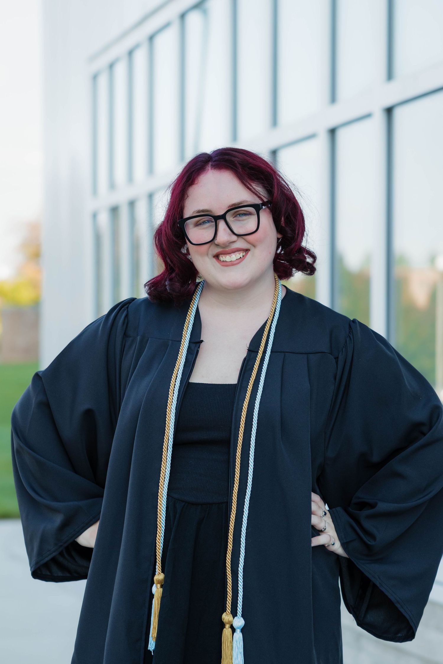 Smiling graduate in black cap and gown with honor cords posing in front of a modern university building.