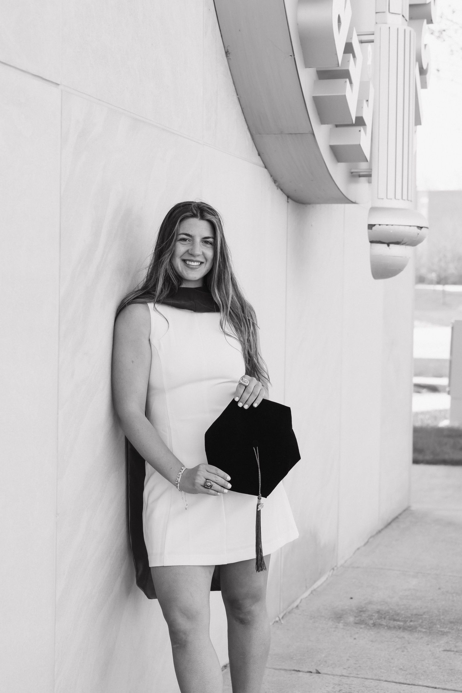 Black and white portrait of smiling female graduate holding mortarboard cap against white wall.