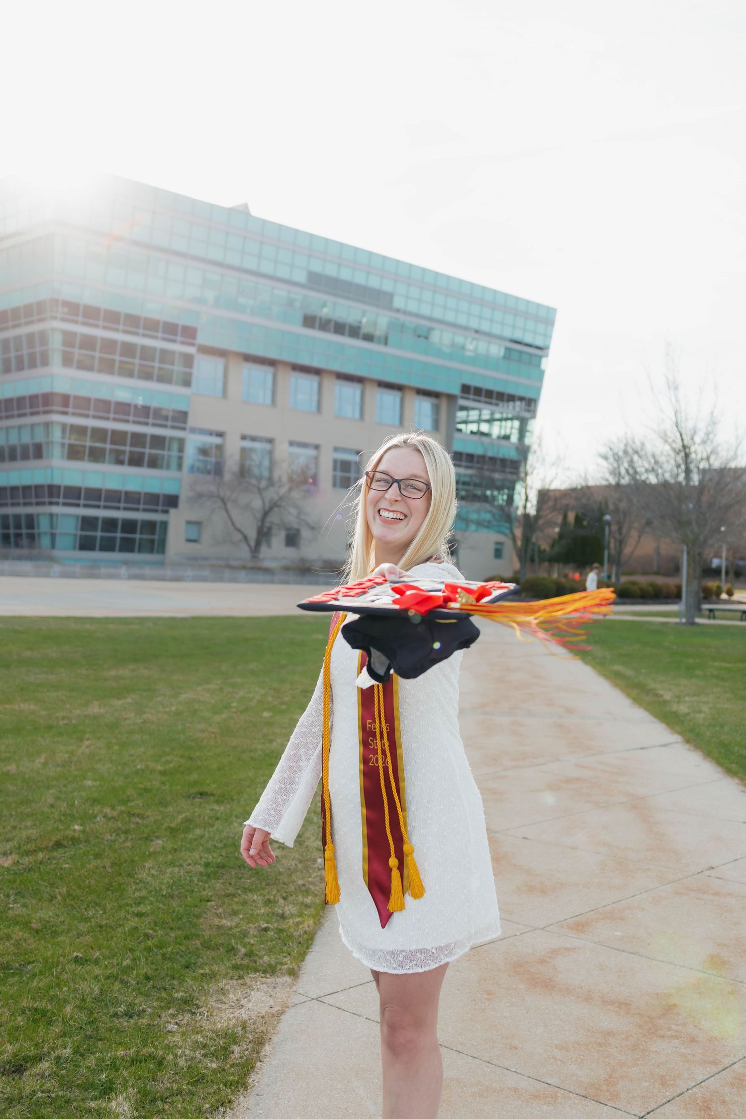 Joyful blonde graduate in white dress tossing cap with colorful stoles on a sunny campus walkway.