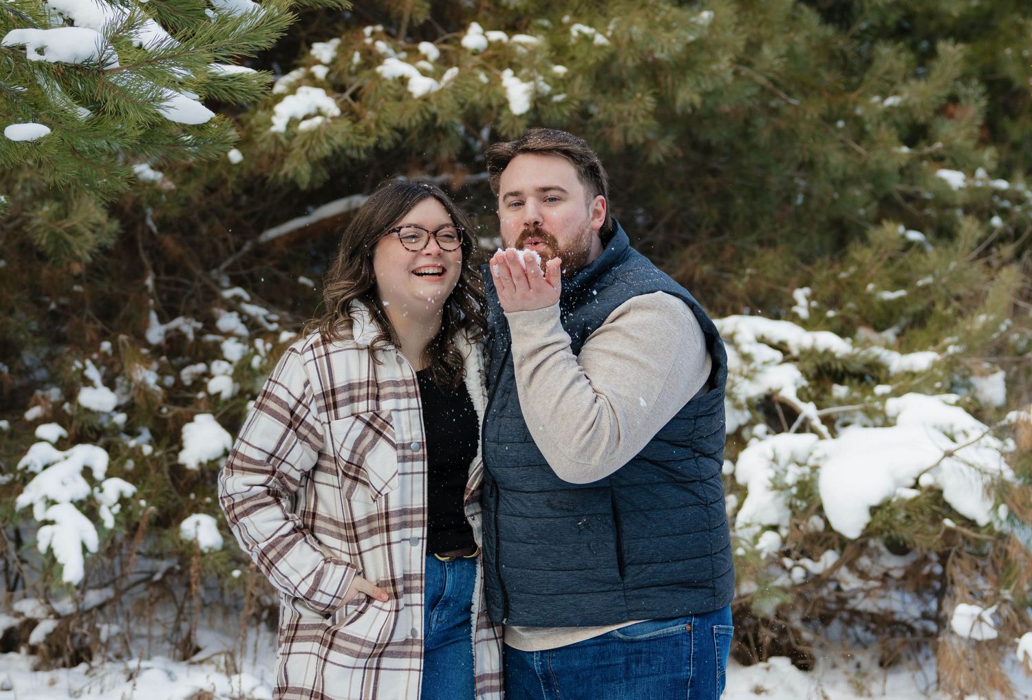 Smiling couple posing playfully in front of snow-covered pine trees, man blowing a kiss to the camera.