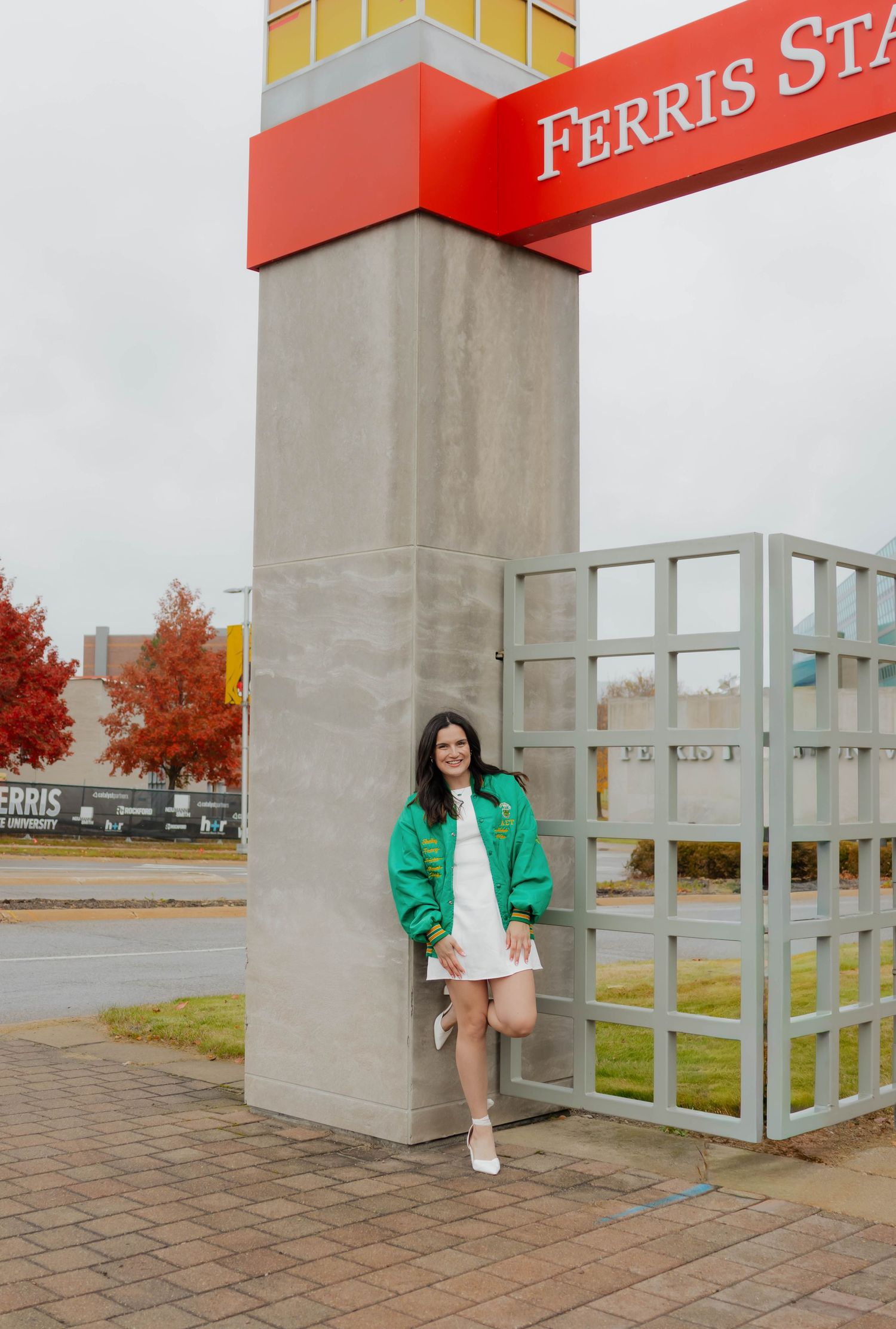 Young woman in green jacket and white dress posing by Ferris State University campus sign.