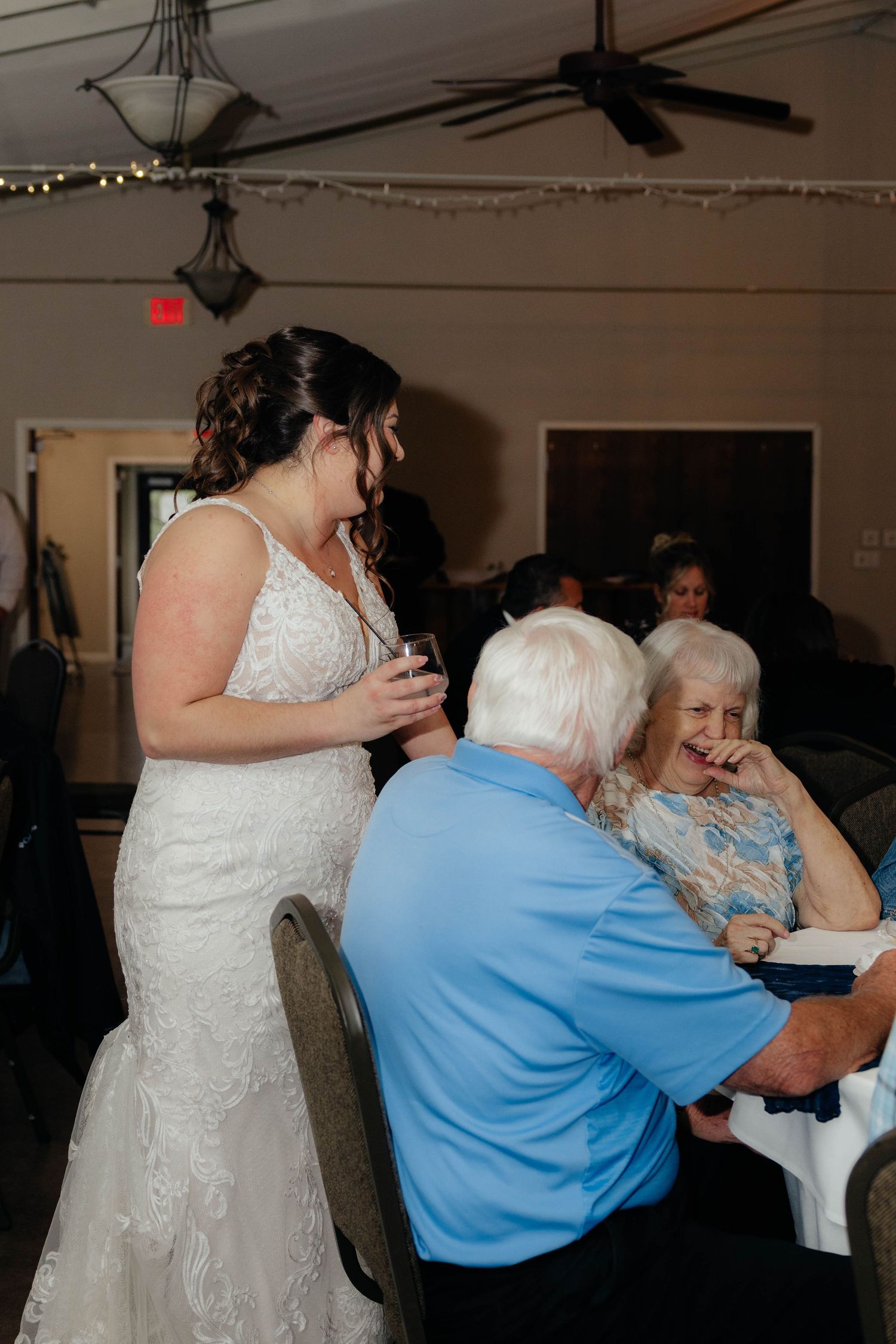 Bride in lace wedding gown laughs and mingles with elderly guests seated at wedding reception table.