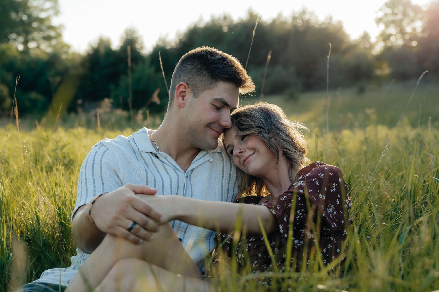 Romantic couple sitting close together in a golden sunlit grassy field, foreheads touching.