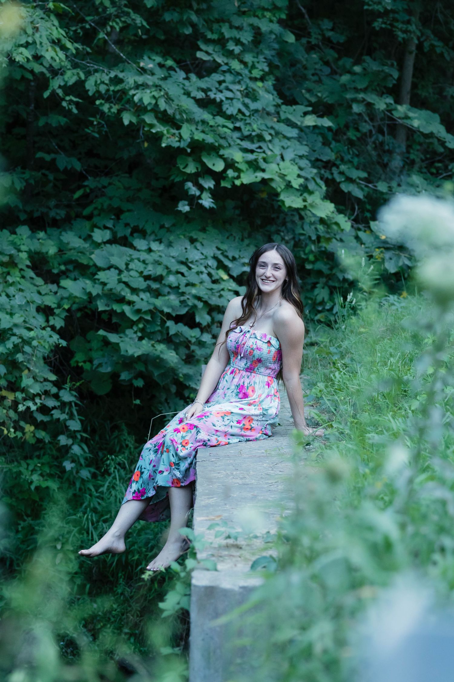 Young woman in floral maxi dress sitting barefoot on stone wall surrounded by lush green forest.