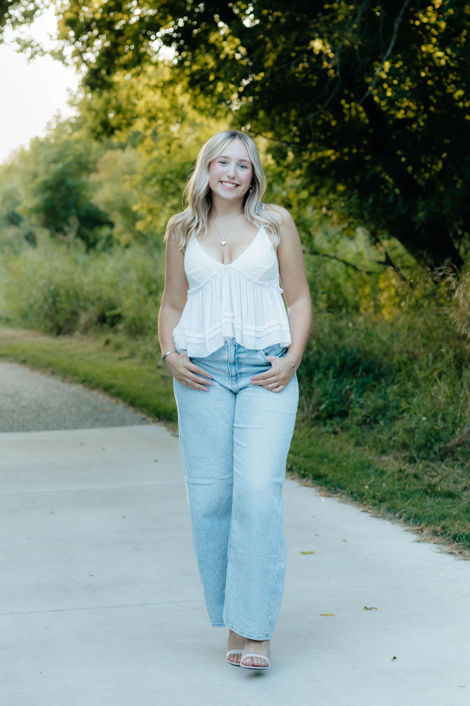 Smiling blonde teen in white cami top and wide-leg jeans walking on park path at golden hour.