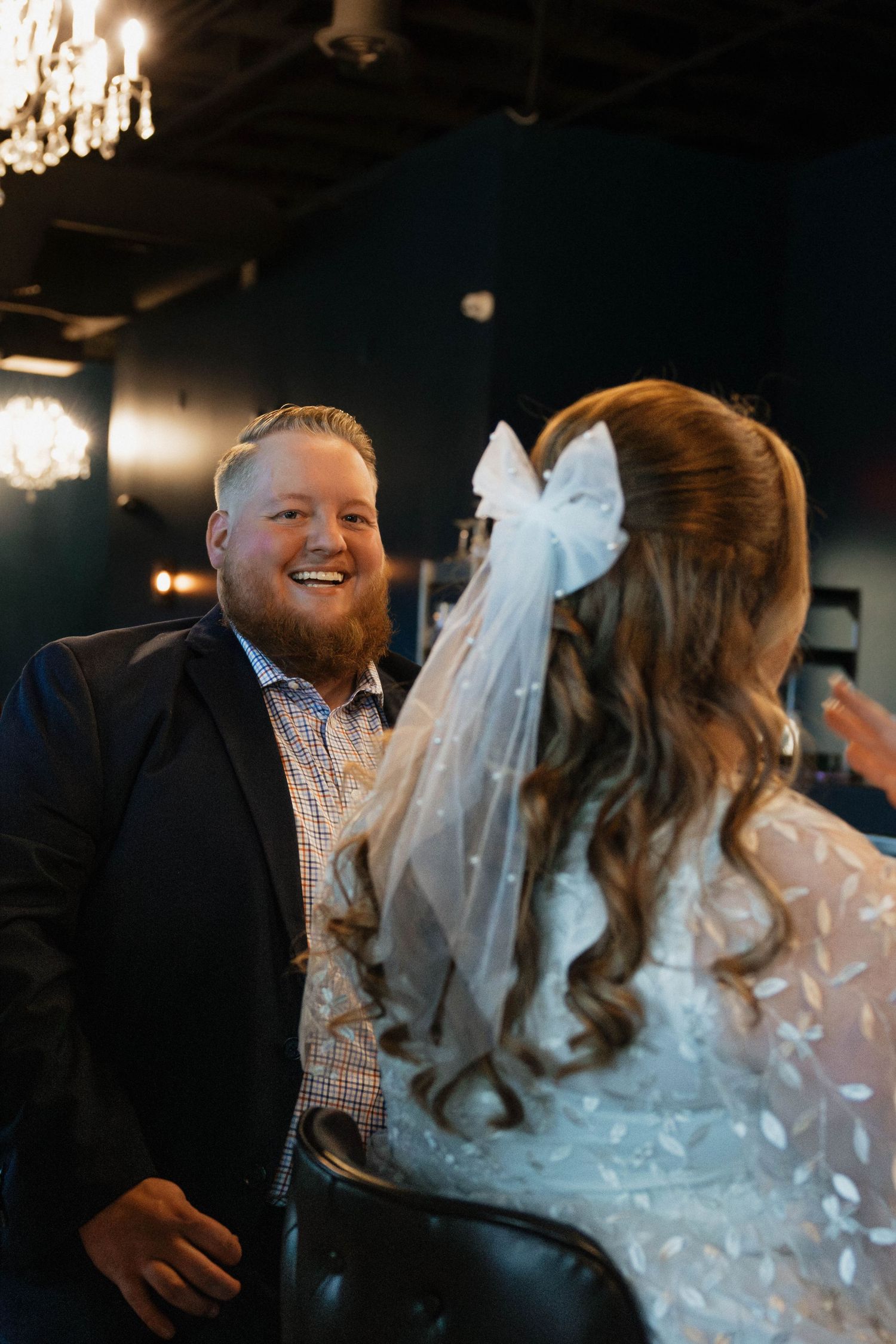 Groom with red beard smiles joyfully at bride wearing a white bow veil indoors.