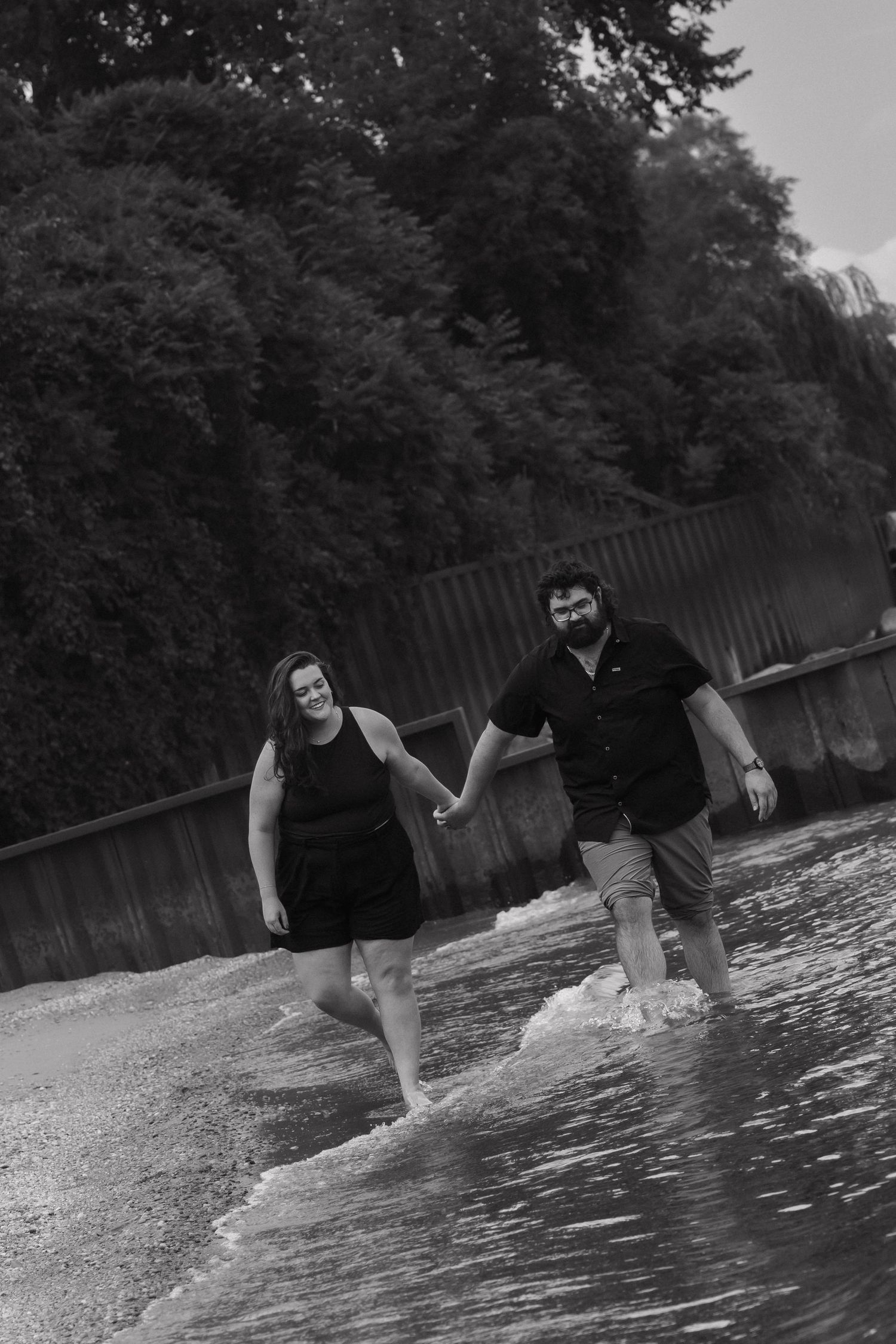 Black and white photo of couple holding hands and wading through shallow water along a tree-lined shore.