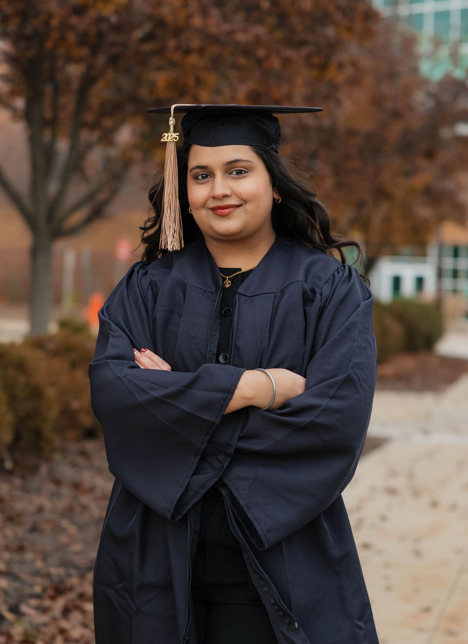 Confident graduate in navy cap and gown posing with arms crossed on a college campus.